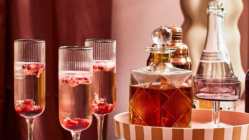 Three champagne flutes filled with pink sparkling wine and red berries, a crystal decanter of amber liquid, a copper cocktail shaker, a bottle of champagne, and a pink coupe glass, all arranged on a striped pink tray against a pink background.