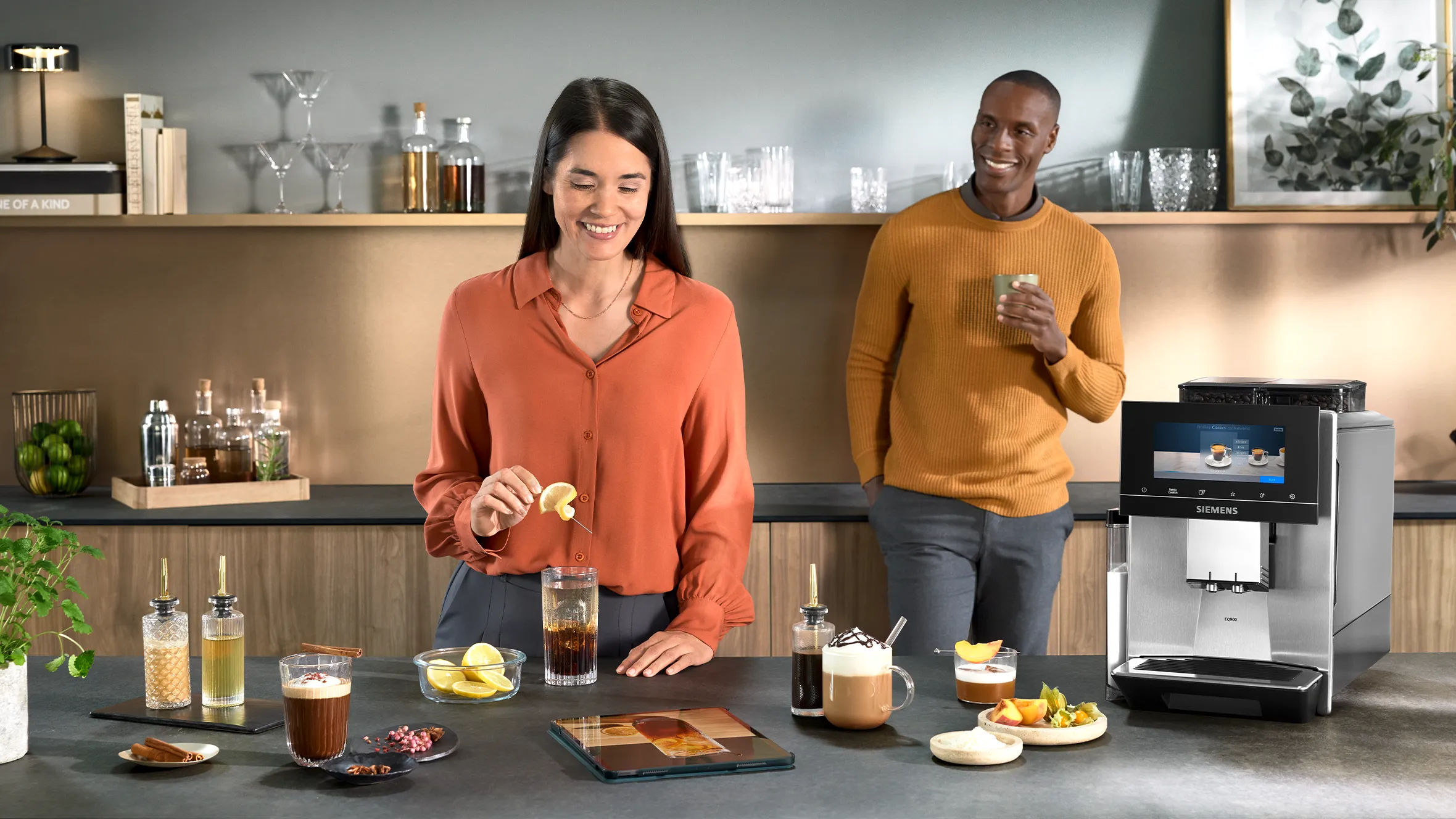 A smiling man and woman stand behind a kitchen counter with a Siemens coffee machine, creating specialty coffee drinks with various syrups and garnishes, guided by a recipe on a tablet.
