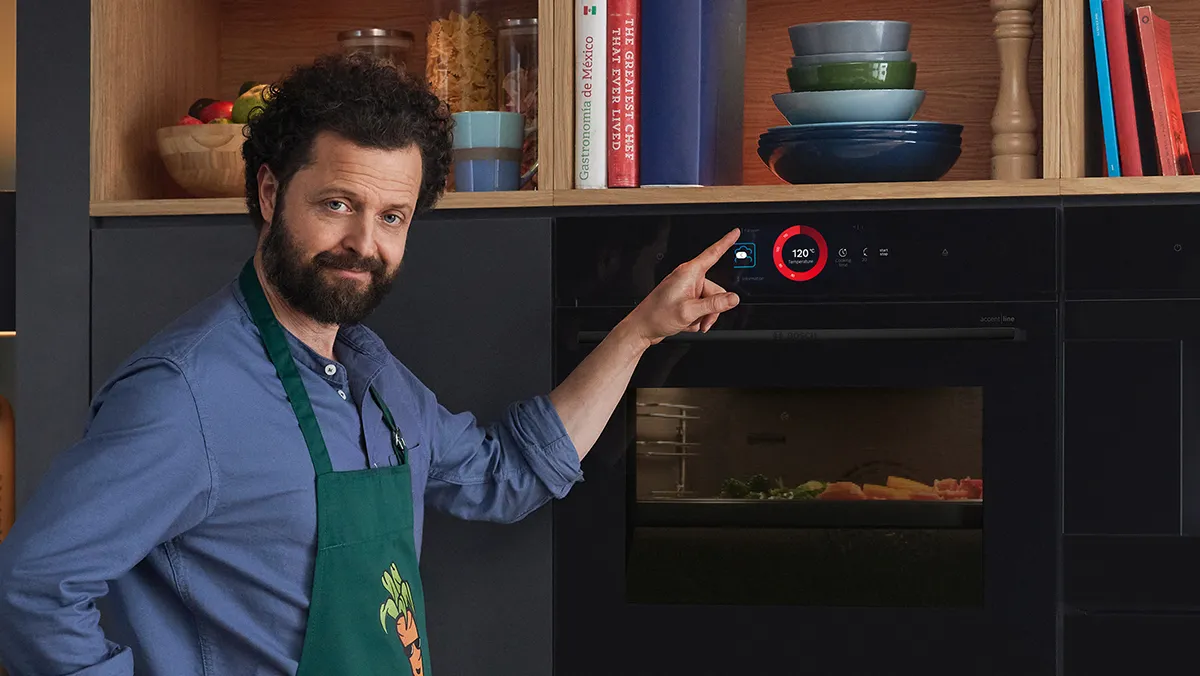 Un uomo con il grembiule è in piedi davanti al forno e indica il pannello di controllo; sullo sfondo ci sono gli scaffali della cucina.
