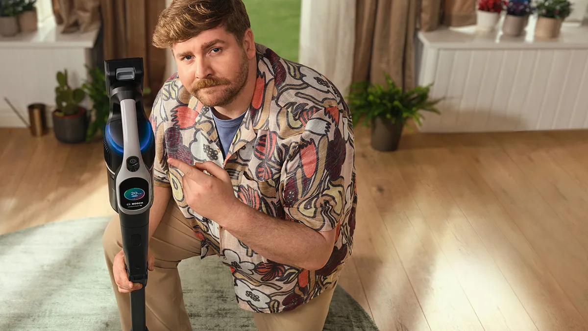 Uomo con barba, camicia con motivi colorati e aspirapolvere in casa.