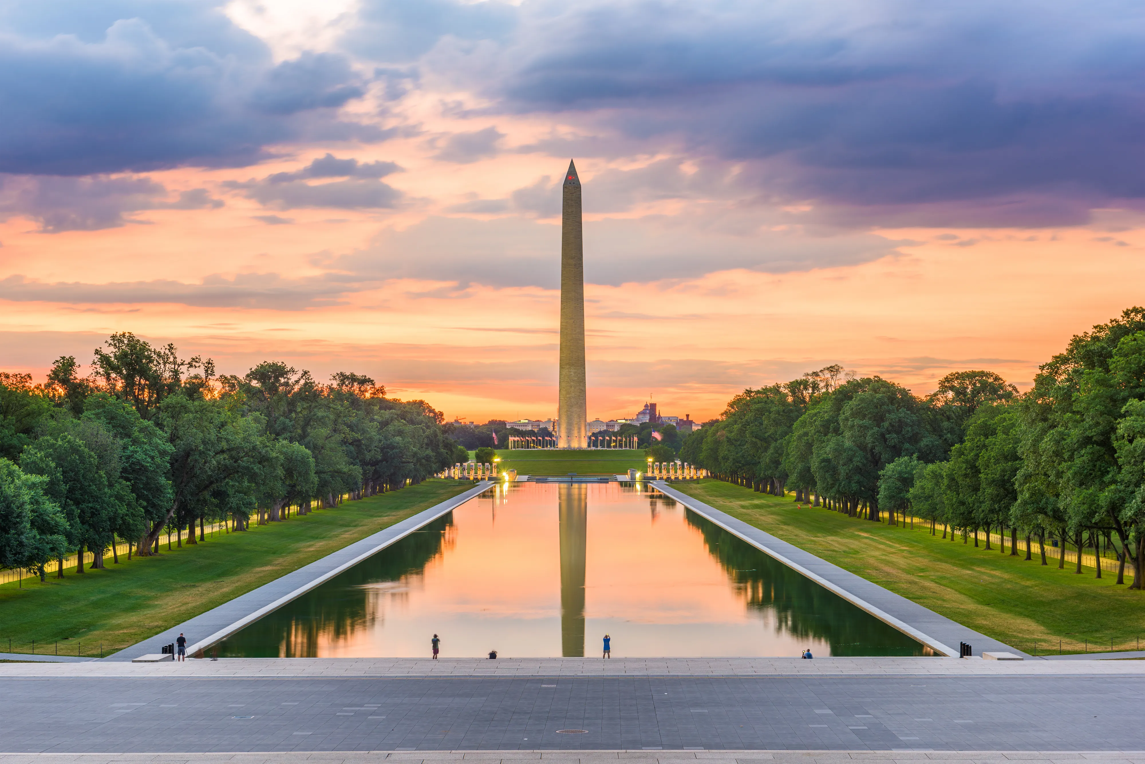 The Washington Monument reflected in the Reflecting Pool at sunset, framed by trees.