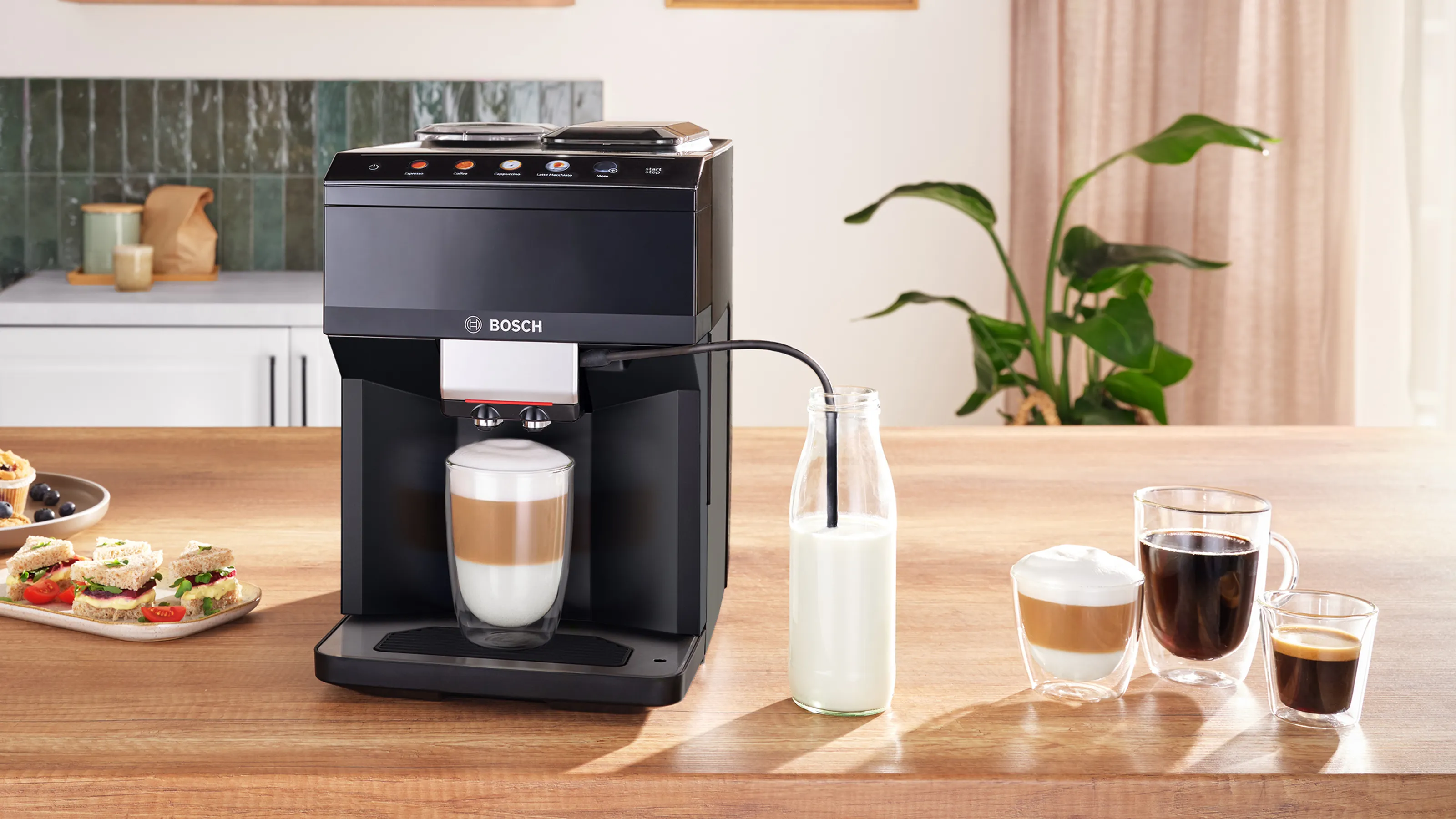 A Bosch coffee machine brewing a latte, surrounded by coffee cups, a bottle of milk, and a plate of sandwiches on a wooden table.