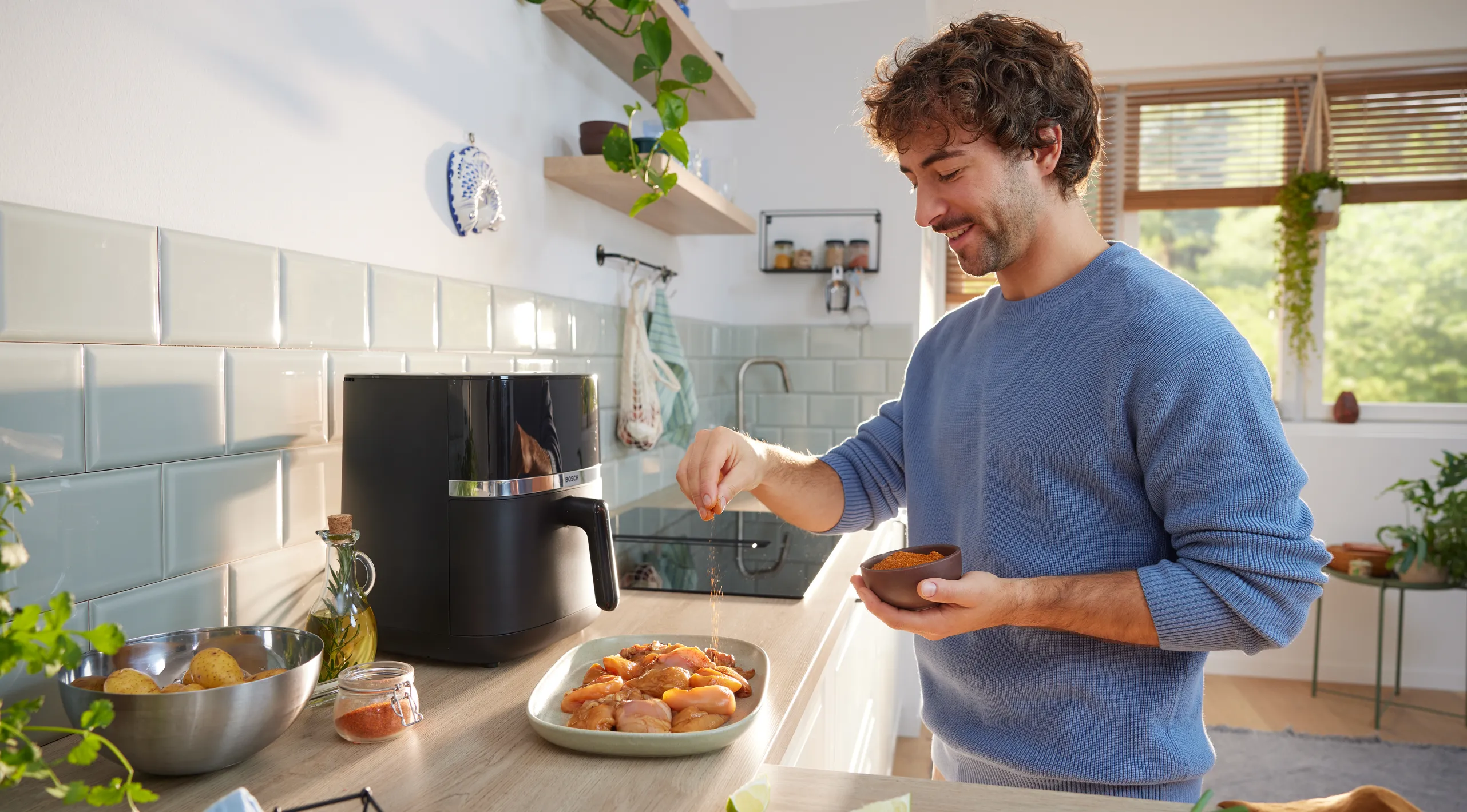 Iemand die rauwe kipstukken op een bord kruidt in een lichte keuken, naast een air fryer en kommen met aardappels en specerijen.