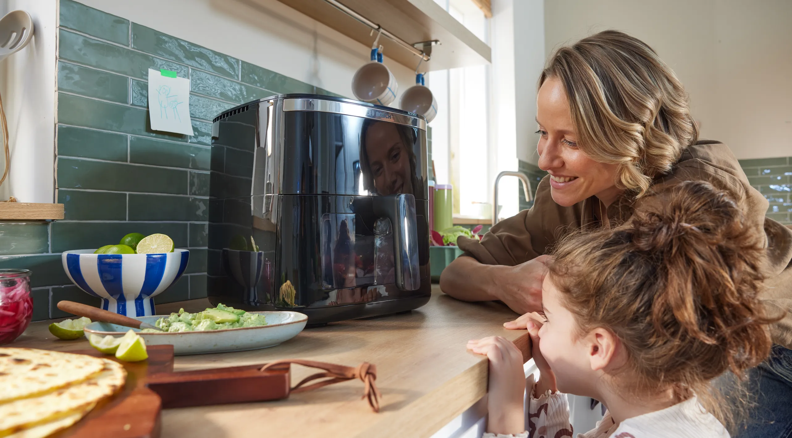 Vrouw en kind kijken naar een zwarte air fryer op het aanrecht, met tortilla's, guacamole en limoenen ernaast.