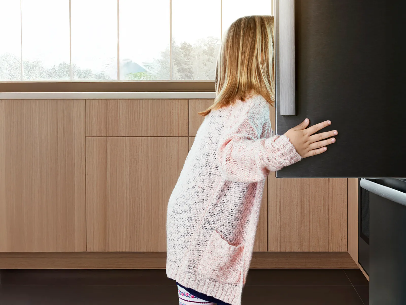 E Chind im rosa Pullover, wo d' dunkle Kühlschranktür in einer Bosch-Küche öffnet.