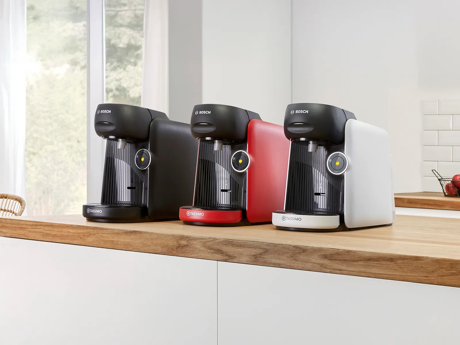 Three Bosch Tassimo coffee machines in black, red, and white, lined up on a wooden kitchen countertop.