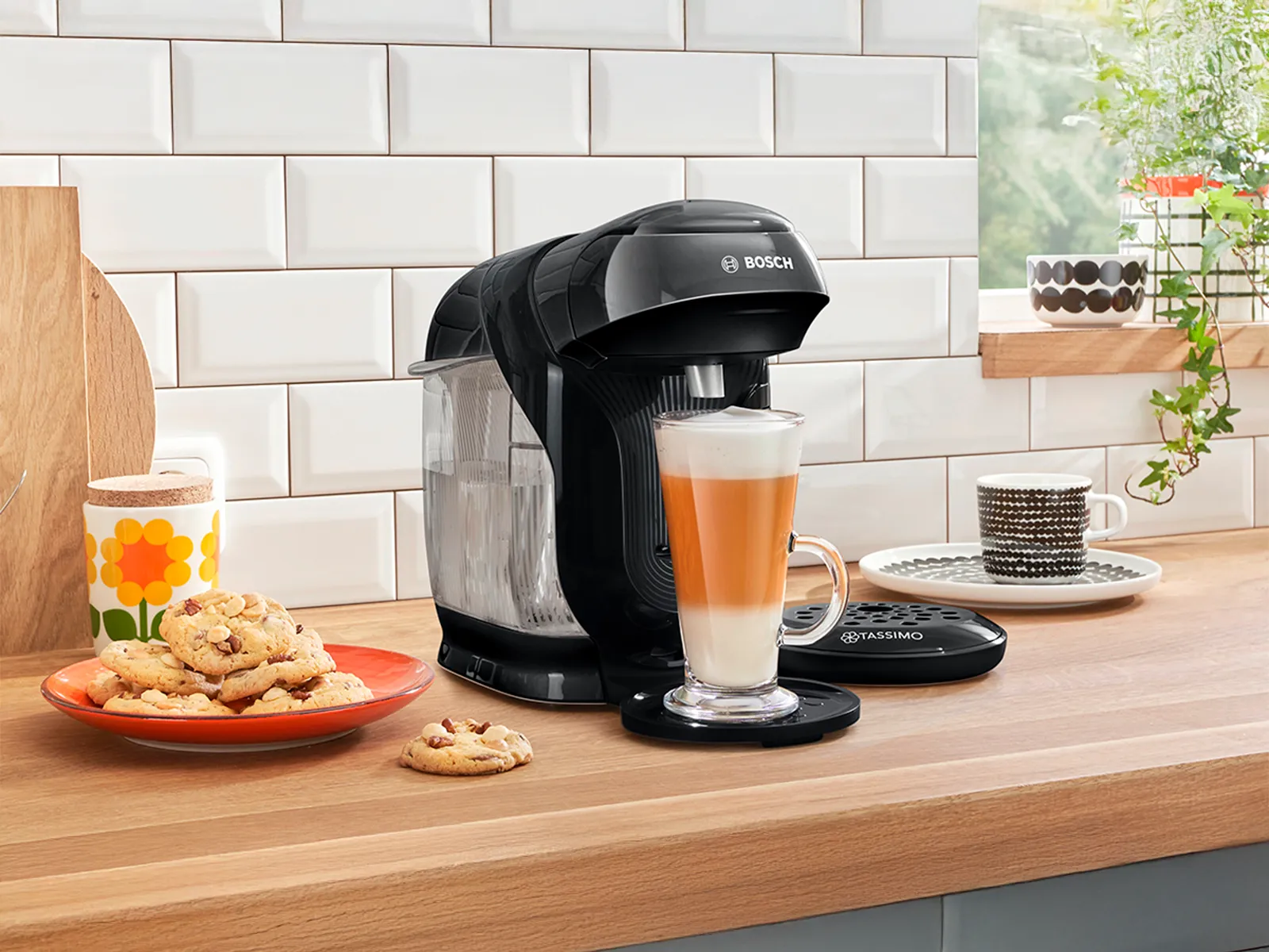 TASSIMO STYLE friendly con un latte macchiato in preparazione sul piano di lavoro di una cucina e accanto un piatto di biscotti.