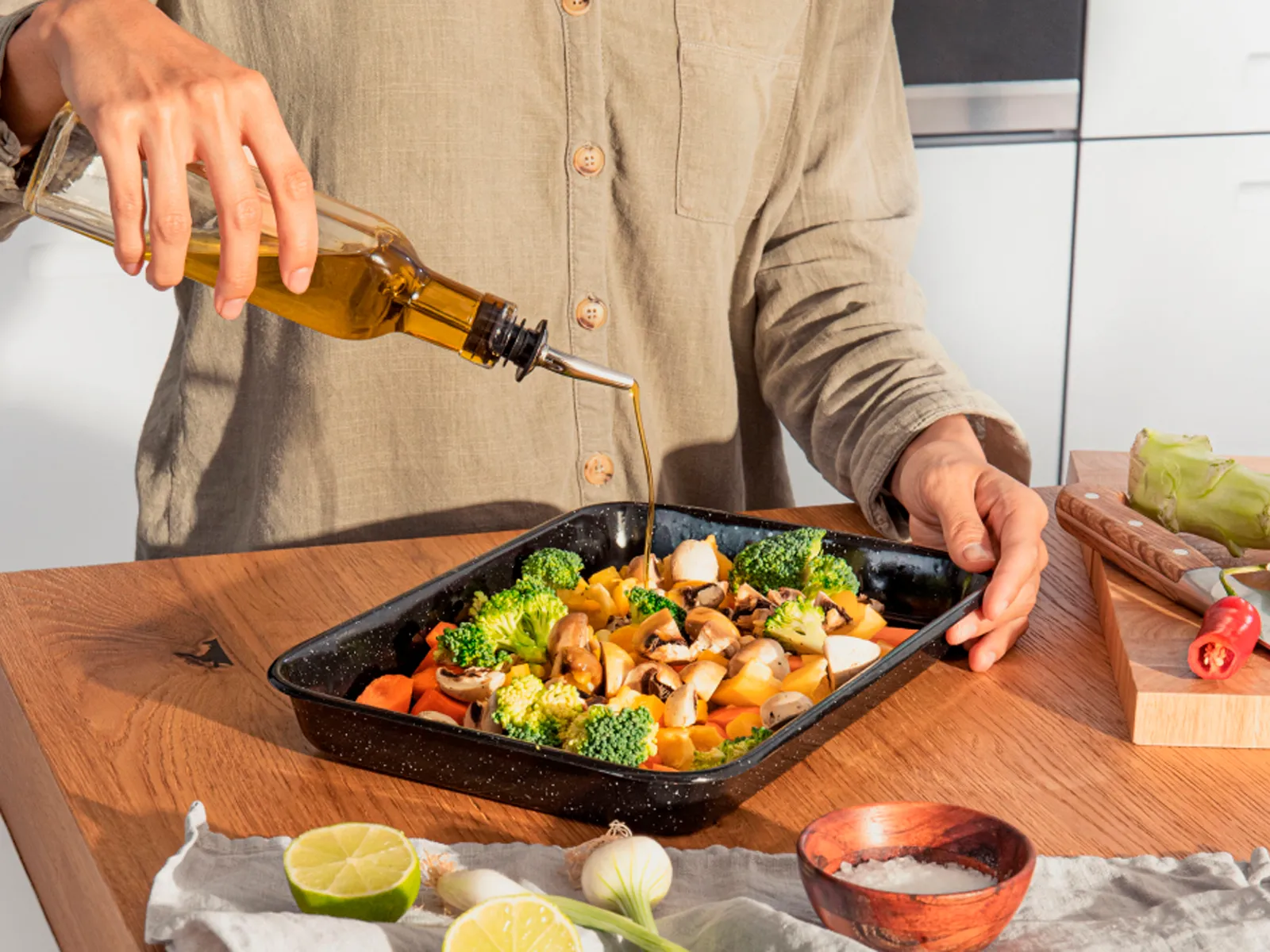 Person drizzling olive oil over fresh vegetables in a baking tray. Preparing a healthy, home-cooked meal with ease.