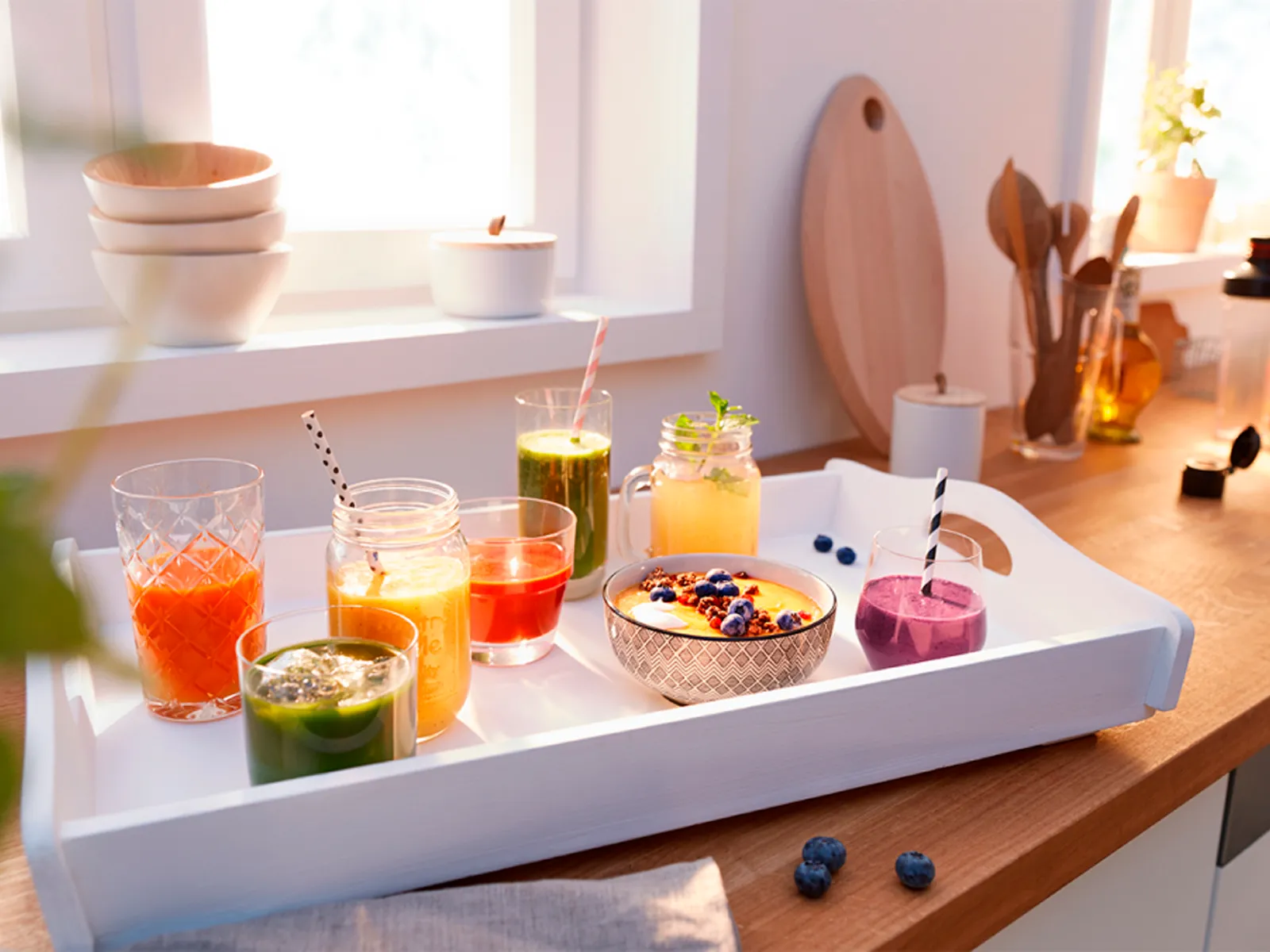 Tray with colorful smoothies and a fruit bowl in a bright kitchen, showcasing fresh, healthy drink options.