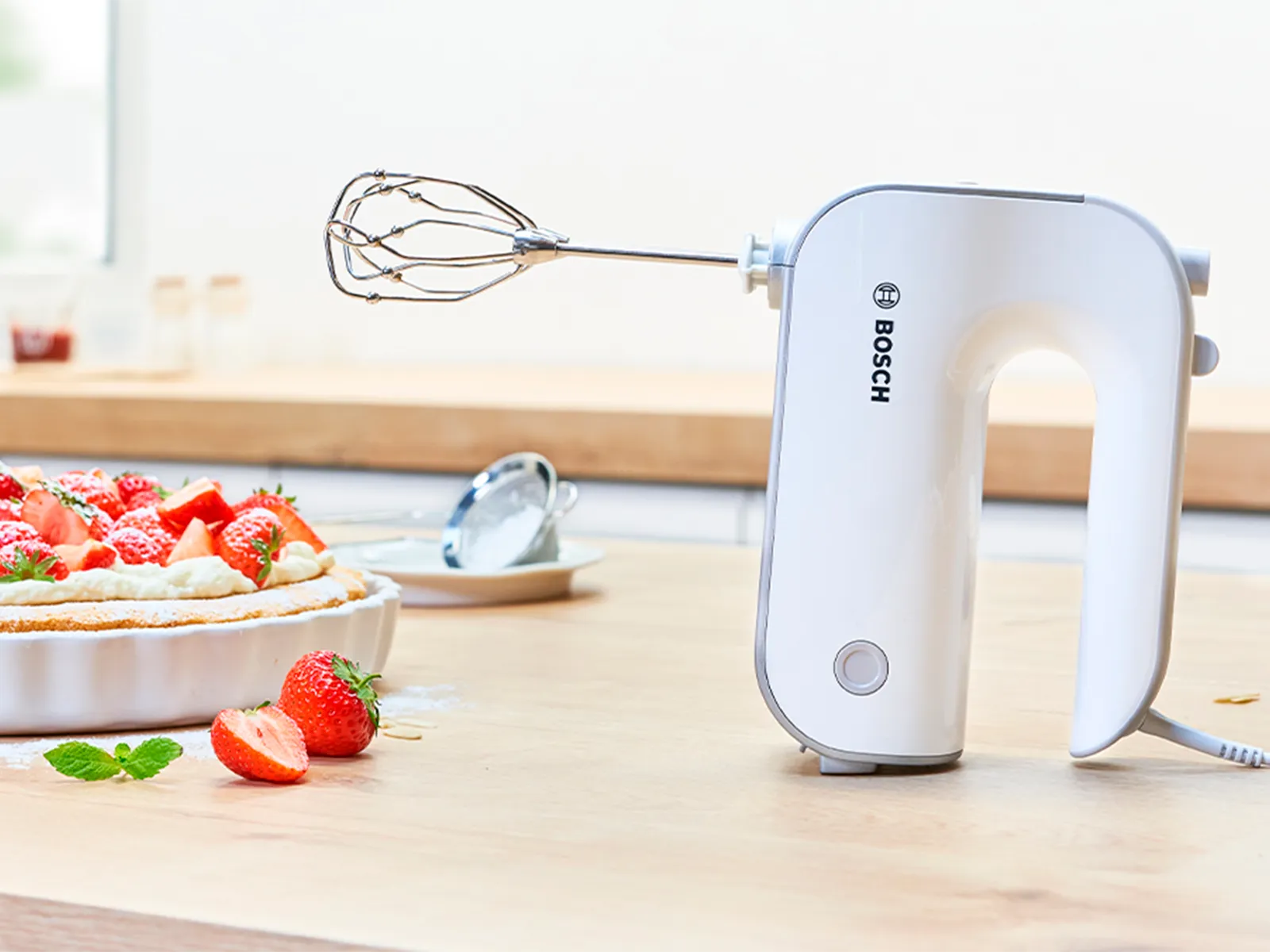 Styline ručni mikser s metlicama na kuhinjskoj radnoj površini, pored šlaga i kolača s malinama.