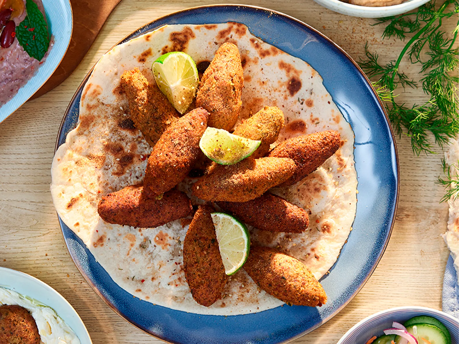 Vedere de sus a unui strat de Kibbeh pe o lipie lângă boluri cu sosuri şi felii de legume.