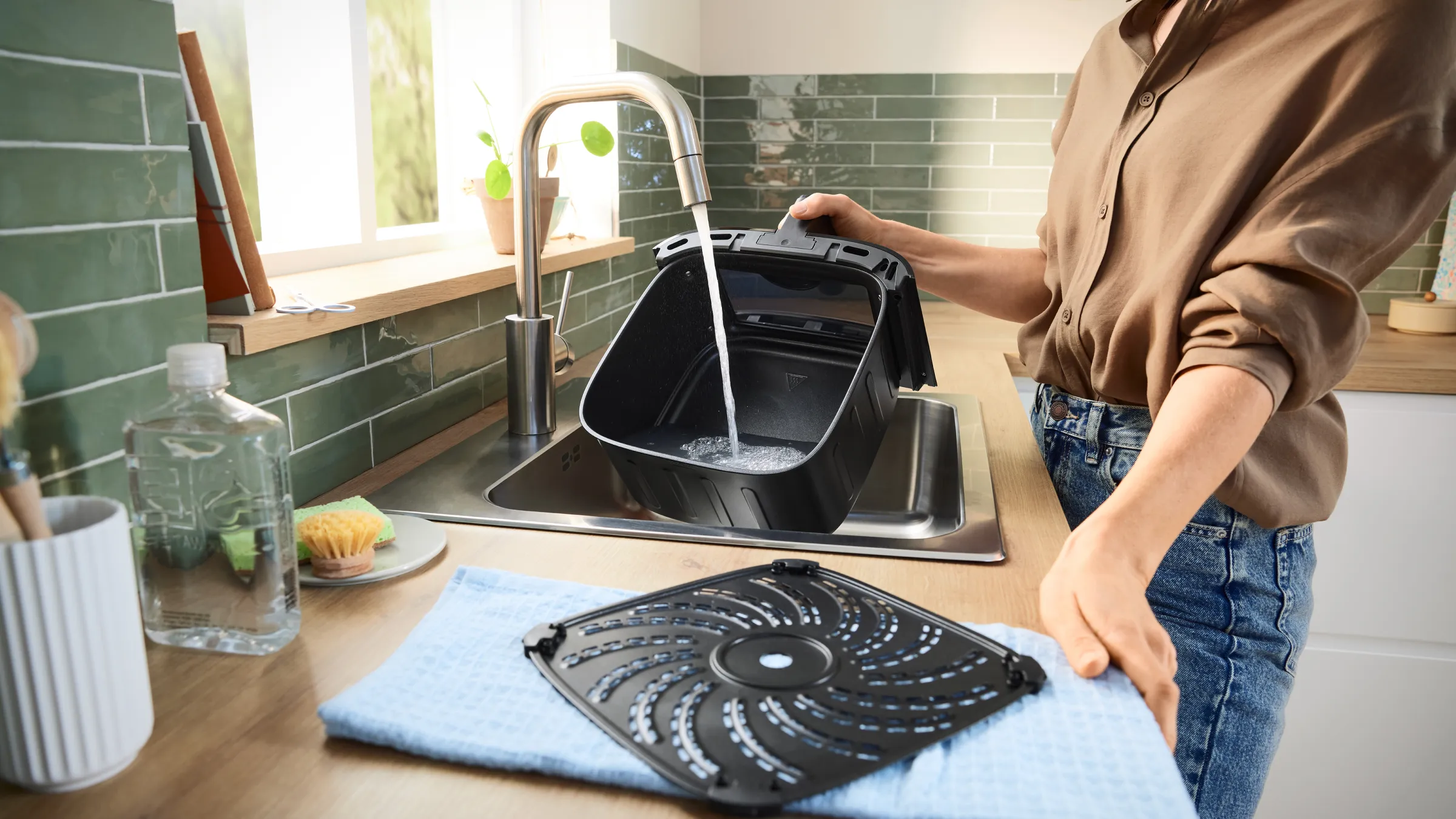 Iemand spoelt een air fryer-bak onder de kraan in een keukenspoelbak; het bijbehorende rooster ligt op een blauwe doek op het aanrecht.