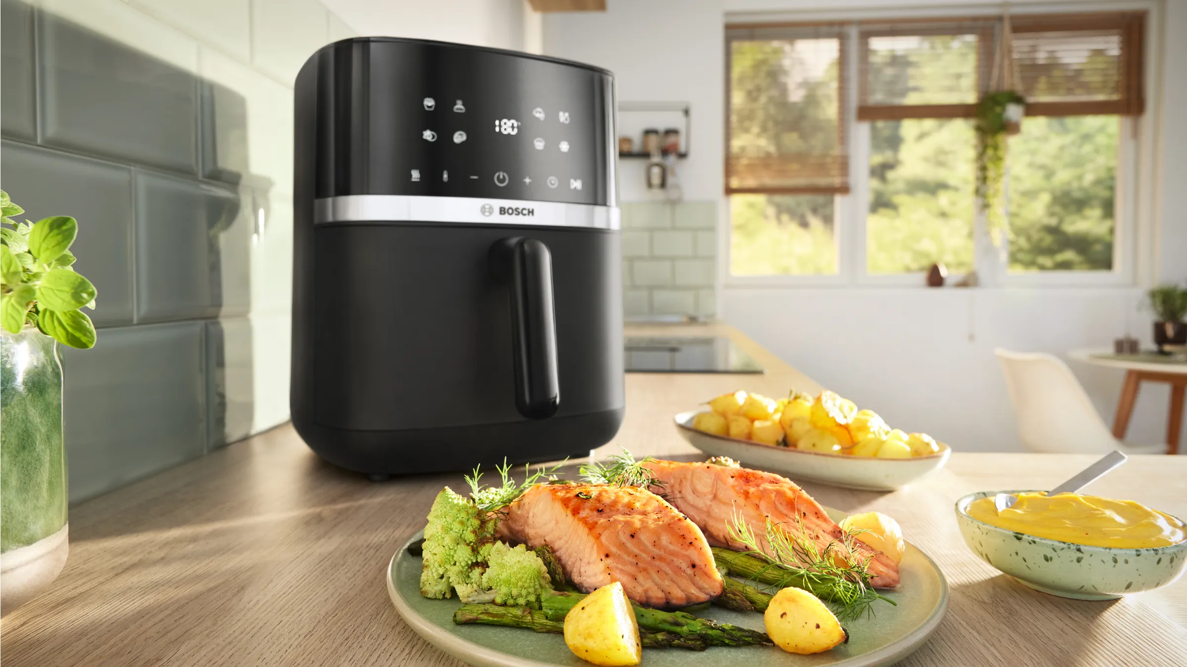 Zwarte Bosch-air fryer op een keukenwerkblad; op de voorgrond een bord met gegrilde zalm, asperges en gebakken aardappeltjes en een kom saus.