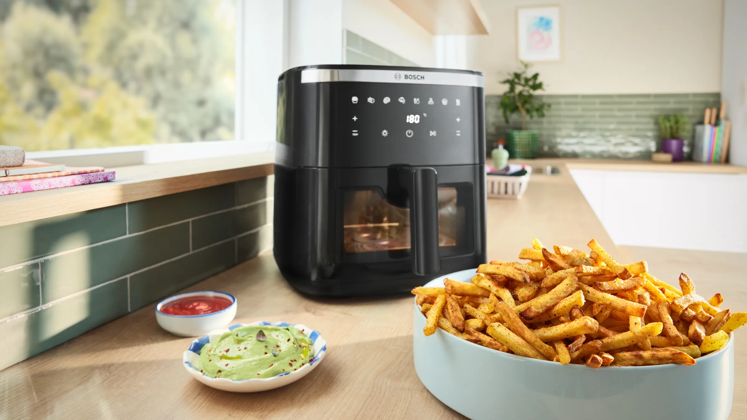 Zwarte air fryer op een licht keukenblad bij het raam, op de voorgrond een schaal met goudbruine frietjes en twee kleine schaaltjes met dips (guacamole en ketchup), met planten en keukengerei op de achtergrond.