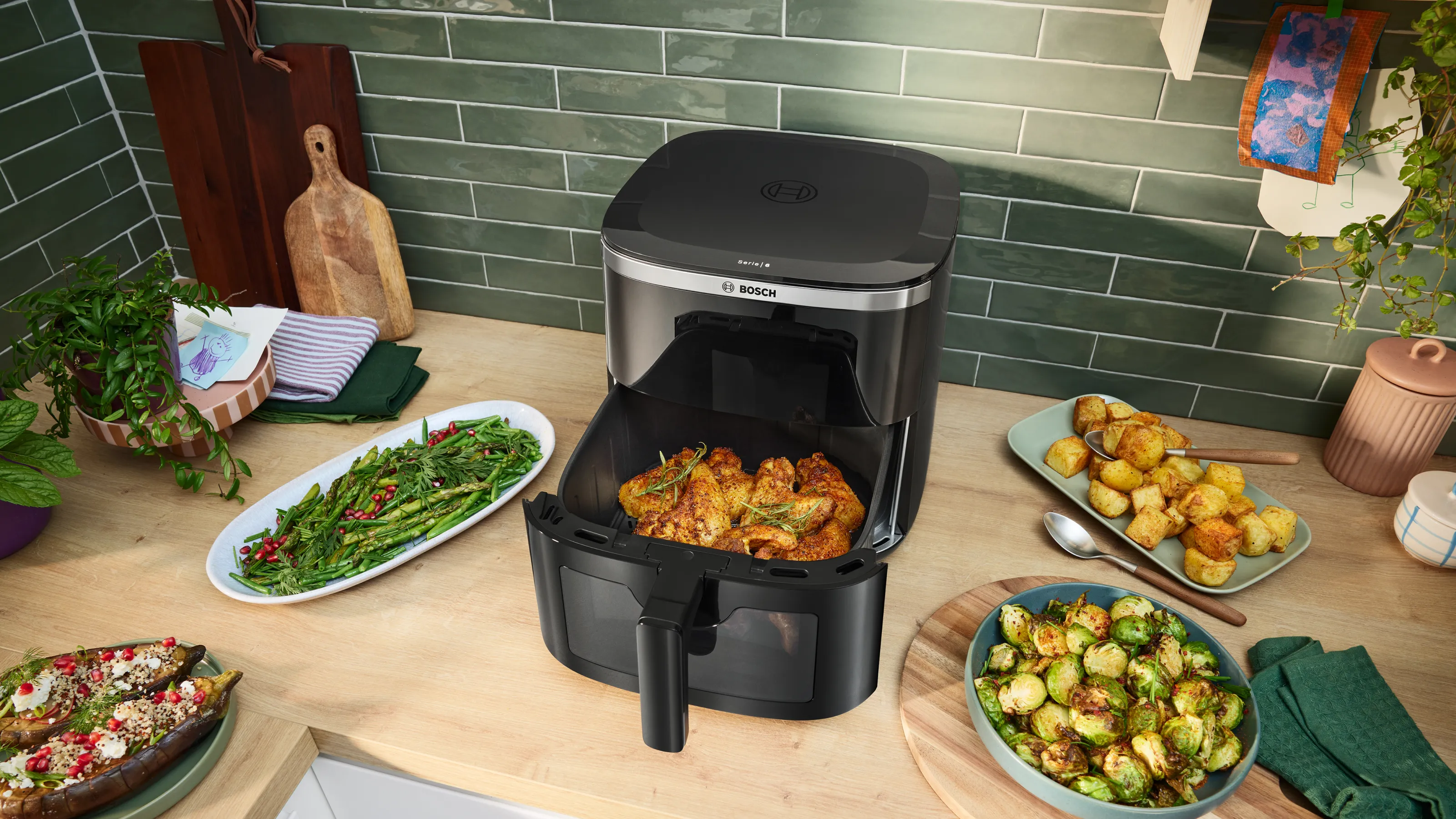 Una freidora de aire de sobremesa llena de alitas de pollo cocinadas, rodeada de platos de verduras asadas, en una cocina moderna.