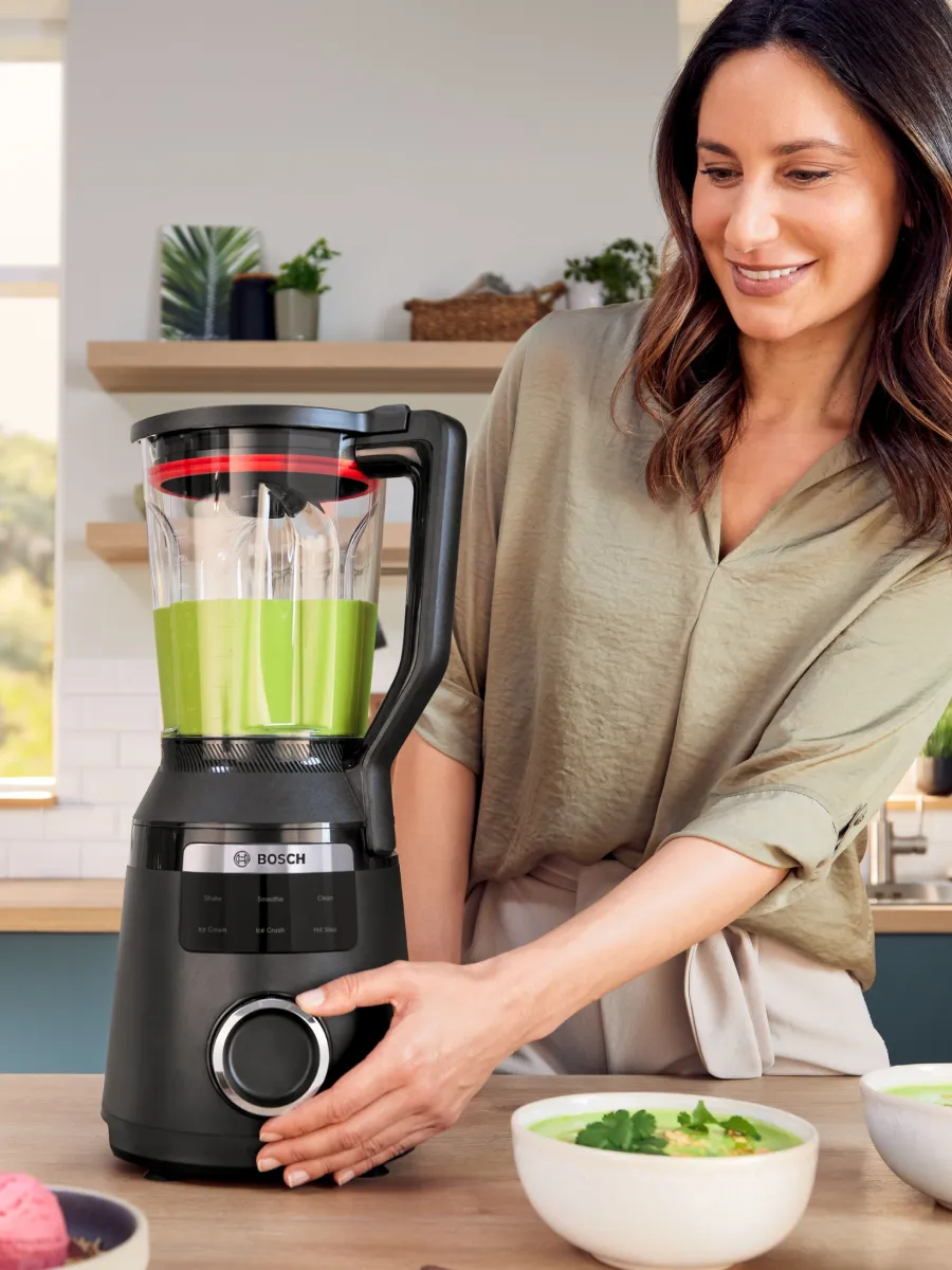 Müasir mətbəxdə bir qadın, Bosch blenderindən istifadə edərək gülümsəyir. Yanında, bir qabda təzə məhsullar var. 