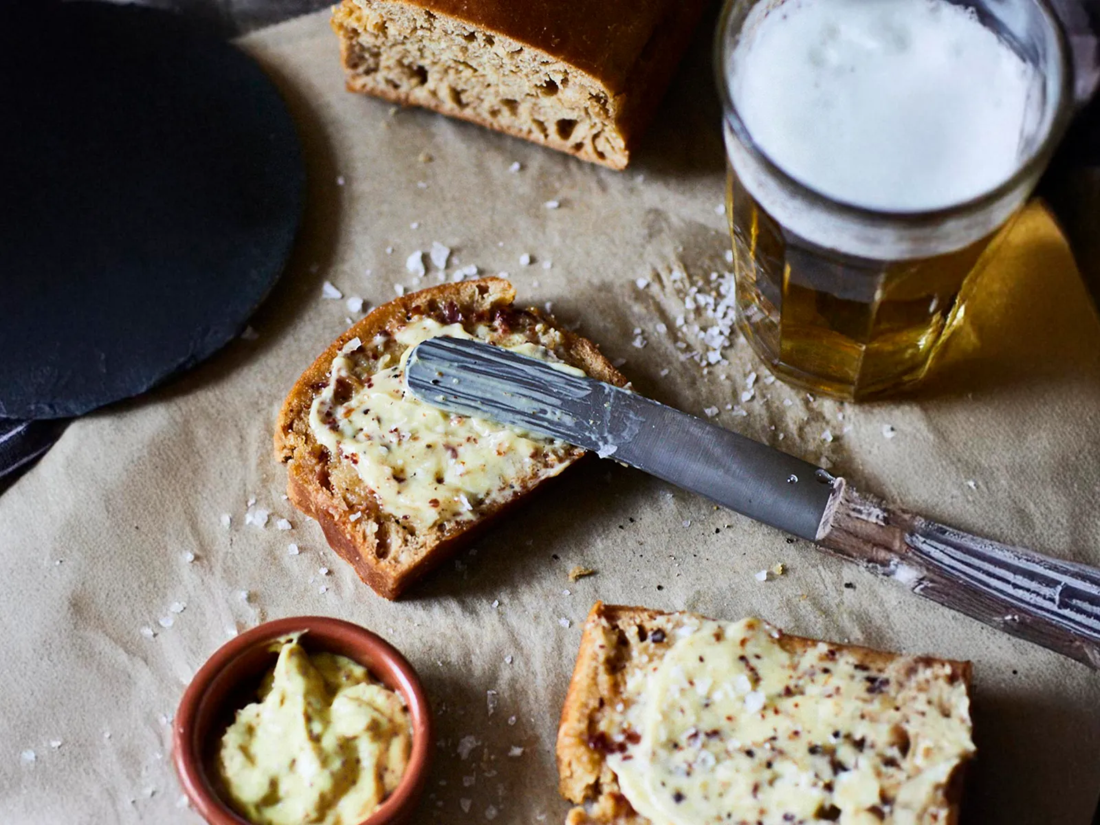 Freshly baked bread with mustard butter spread, a butter knife, flaky salt, and a glass of beer on parchment paper.