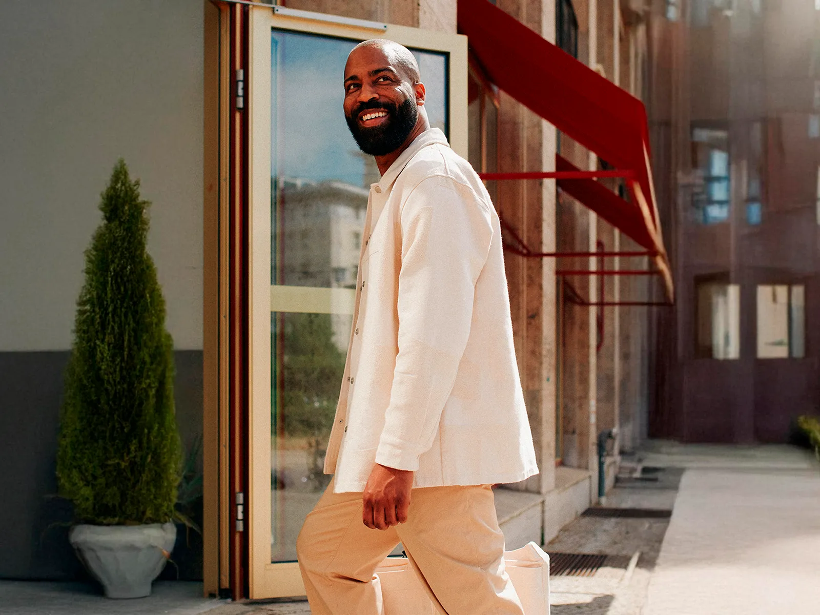 Smiling man in a light-coloured outfit walking outside a modern building with glass doors.