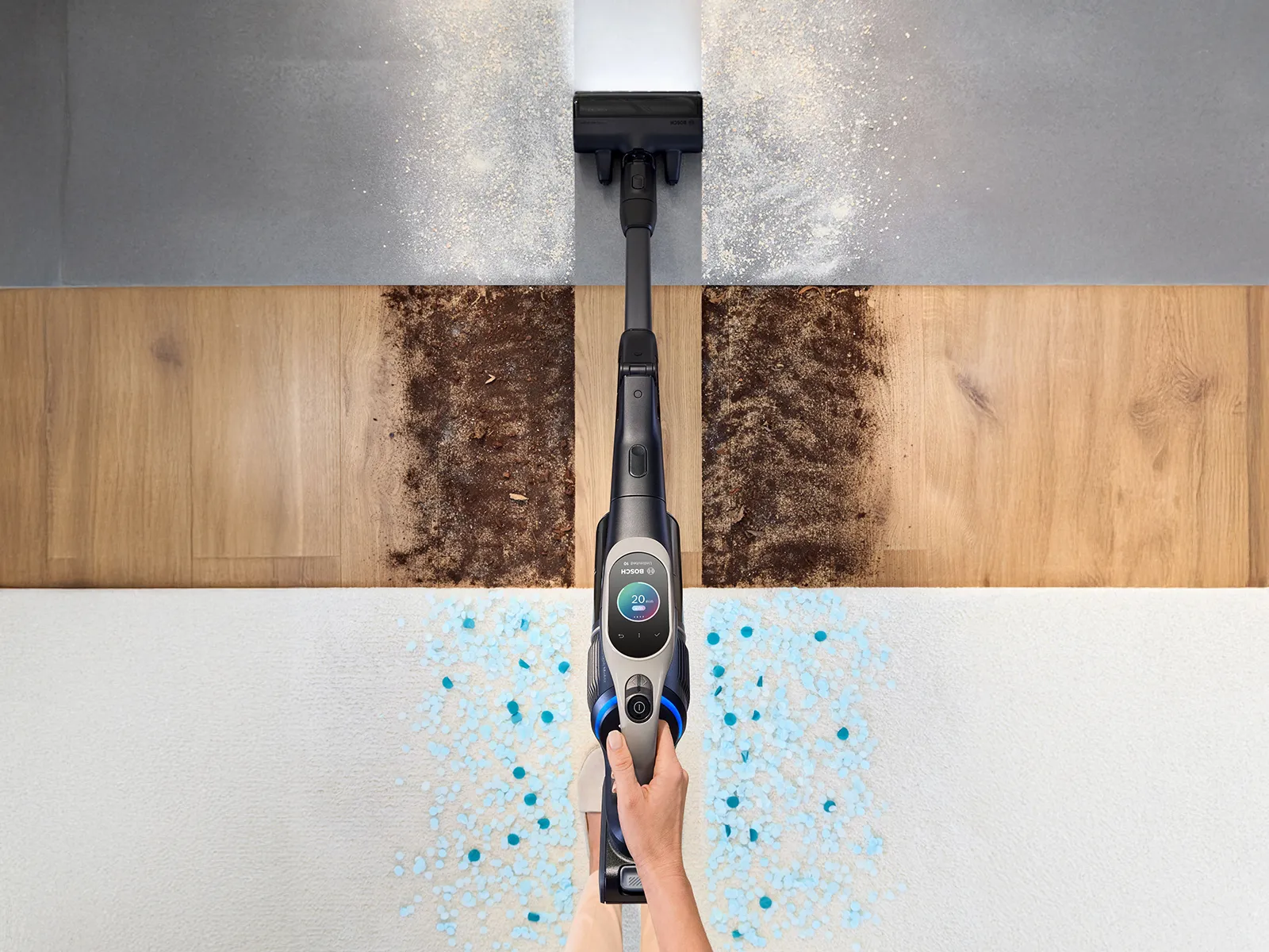 Záběr z vysokého úhlu na bezdrátový vysavač, který projíždí přes tři různé povrchy pokryté špínou a jinými suchými nečistotami, přičemž zanechává za sebou dokonale čistou stopu.