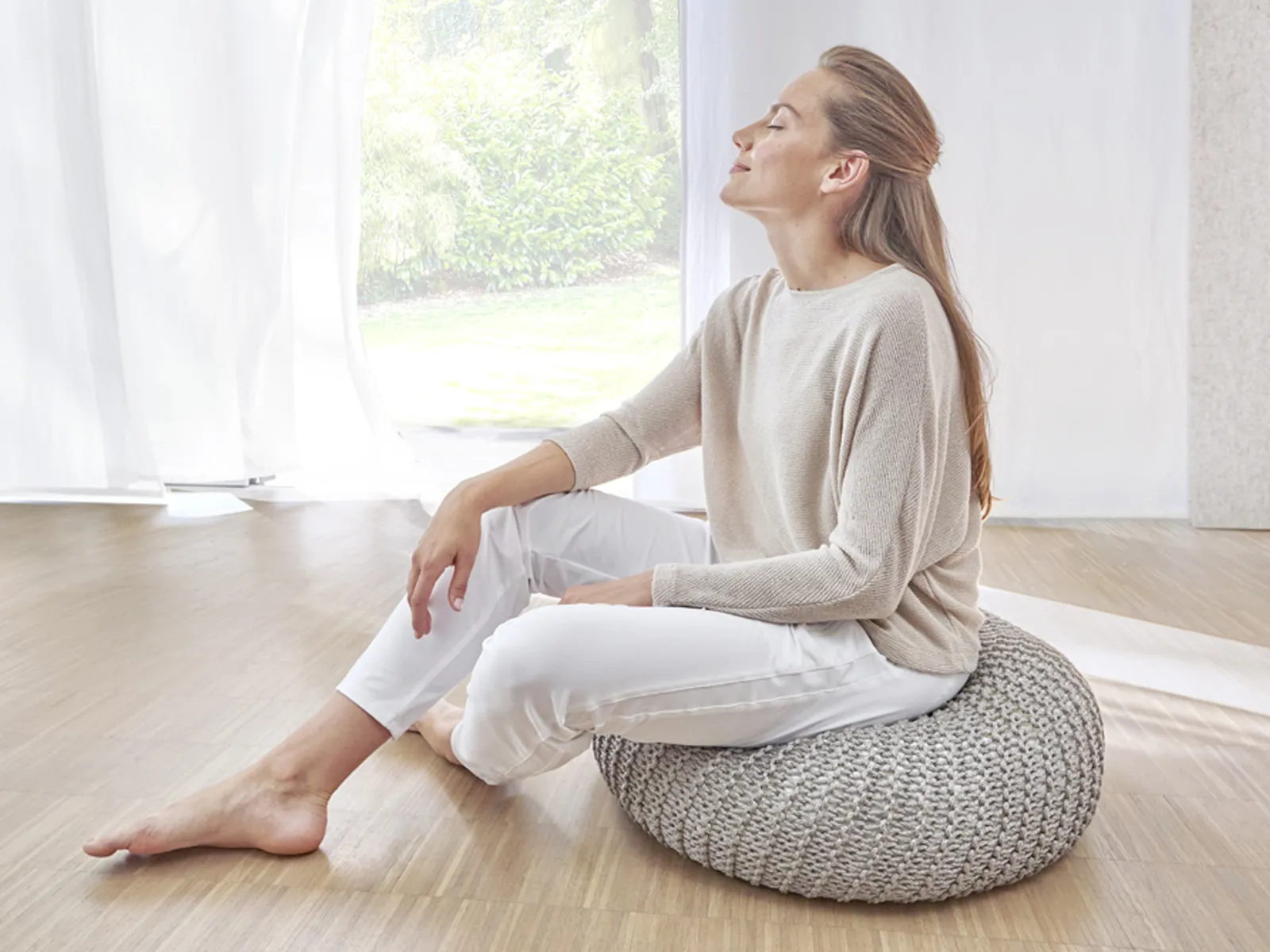 A woman sits on a pouf in the living room and takes a deep breath.