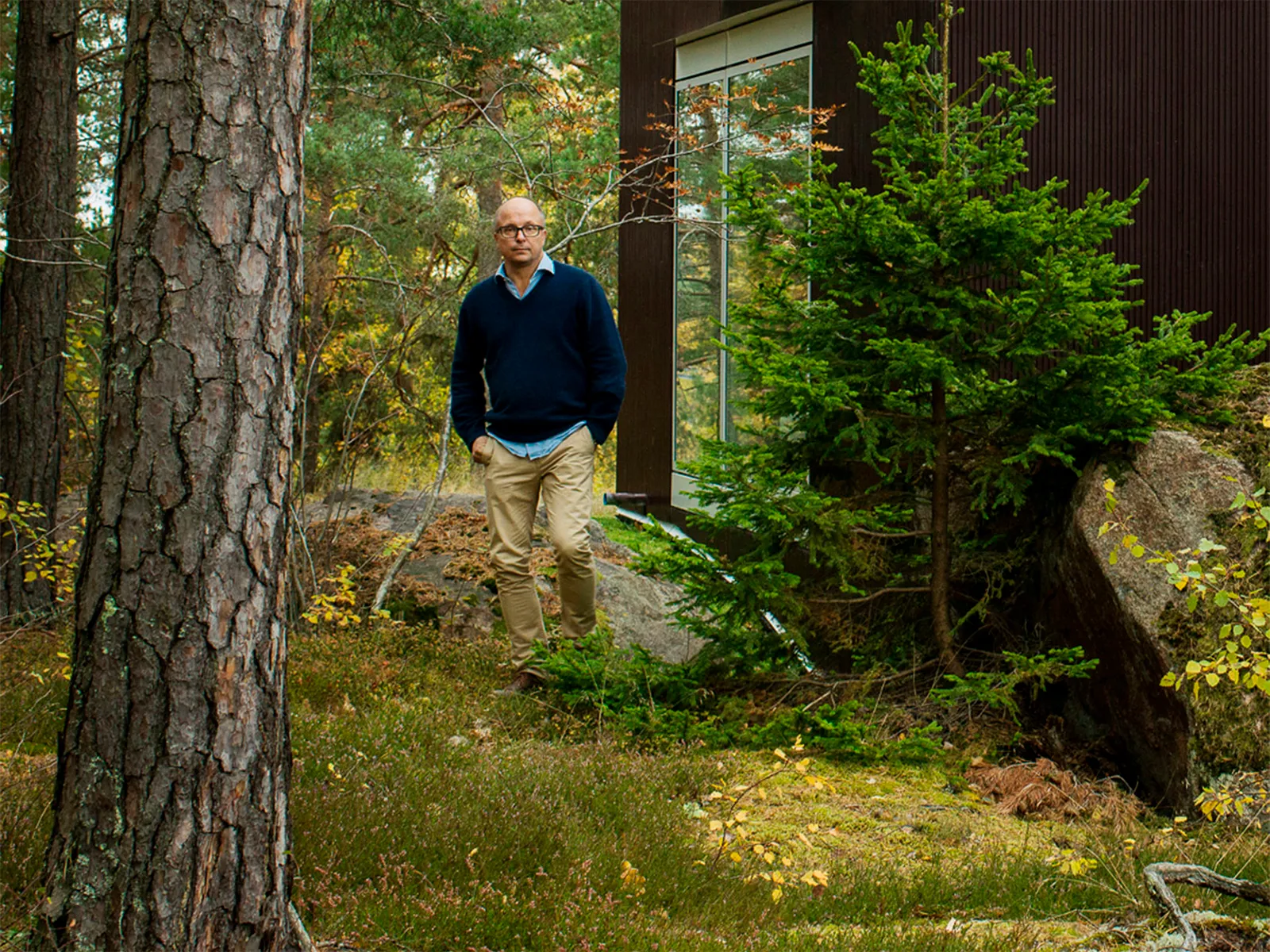 Man in a navy sweater and beige trousers walking through a forested area near a modern house with large windows.
