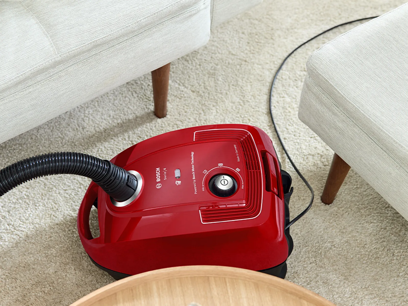 A compact, red Bosch bagged cylinder vacuum manoeuvres between two couches and a table.