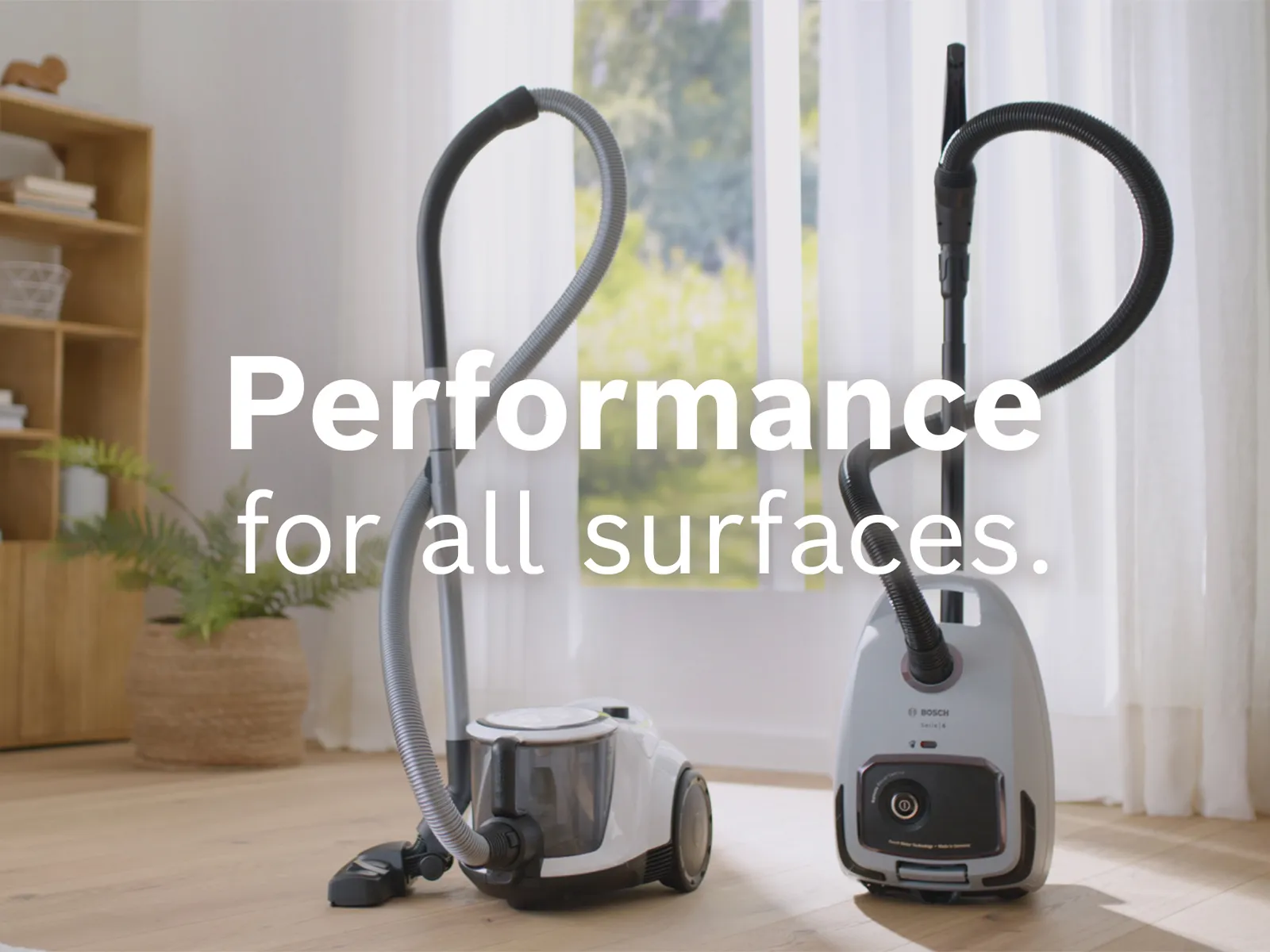 Two Bosch bagless cylinder vacuums stand in front of a window in a study with the words “performance for all surfaces” superimposed over them.