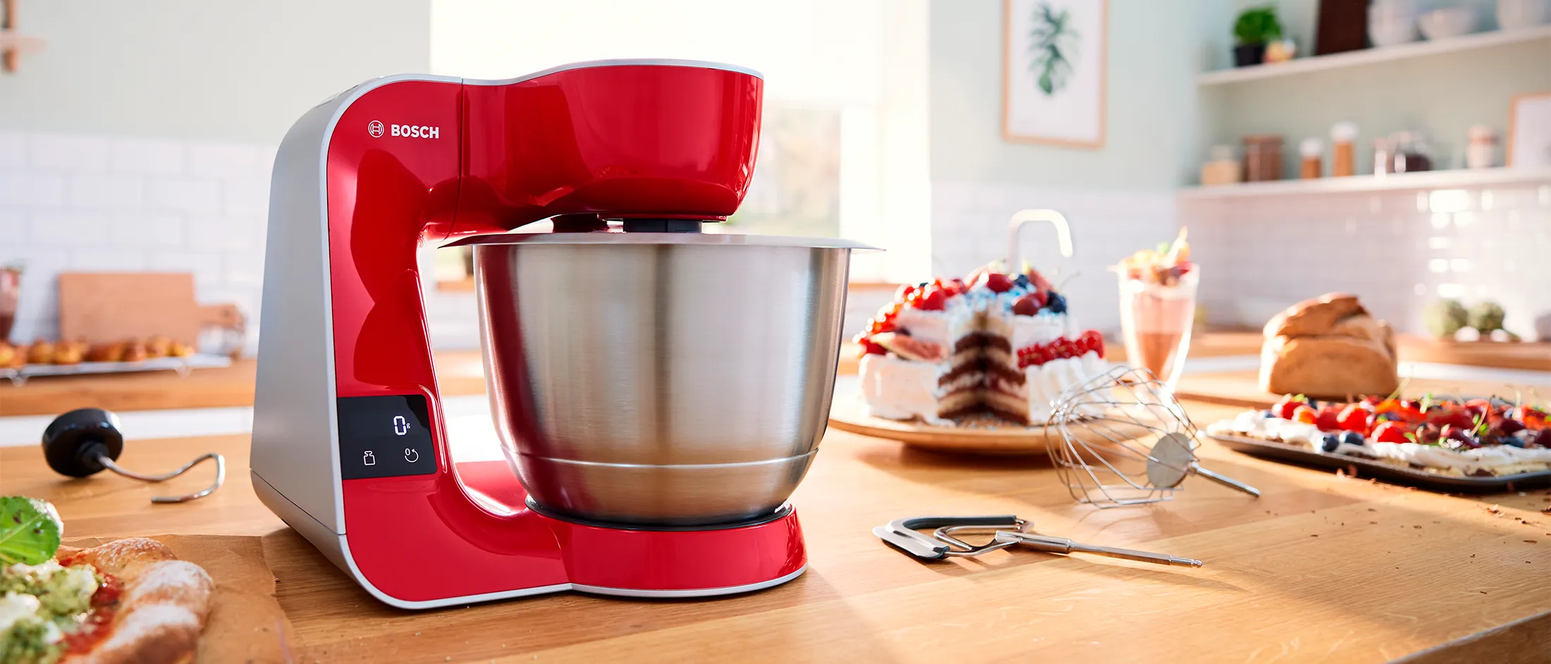 A Bosch Series 4 stand mixer in red surrounded by variaus dishes, like pizza, cake and sweet deserts.