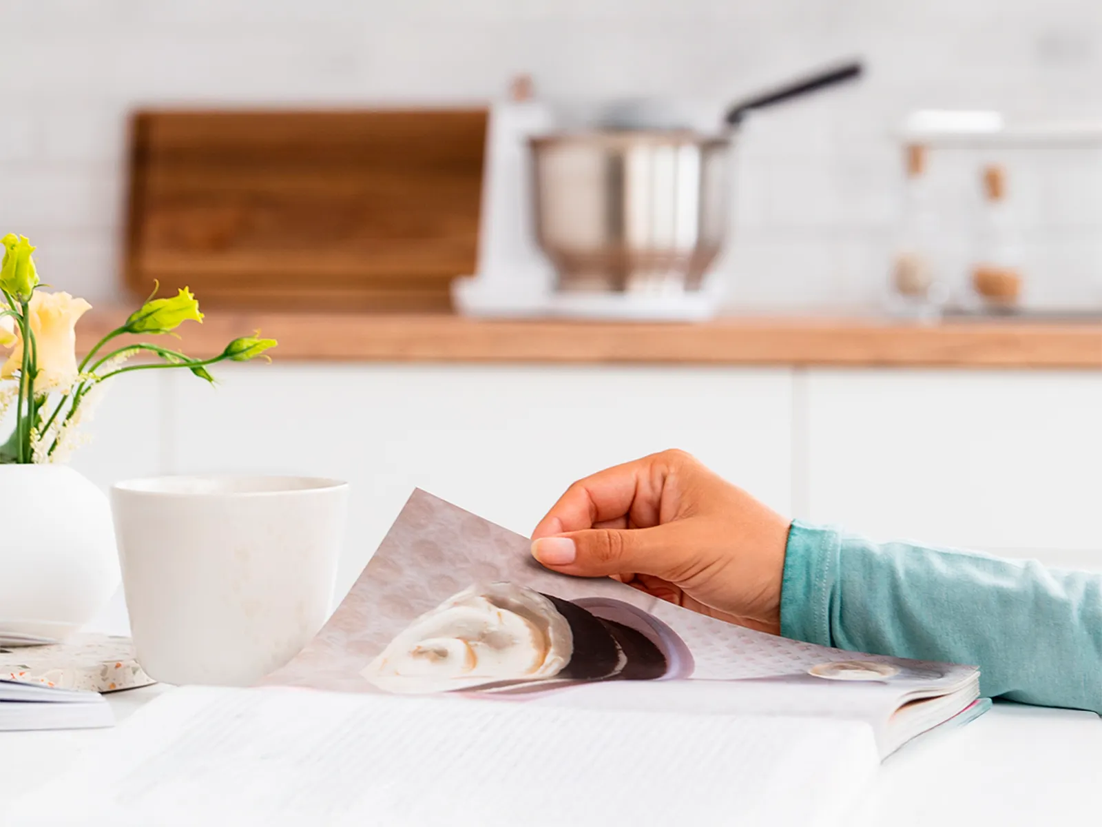 Käsi esitteineen, taustalla keittiövälineitä.