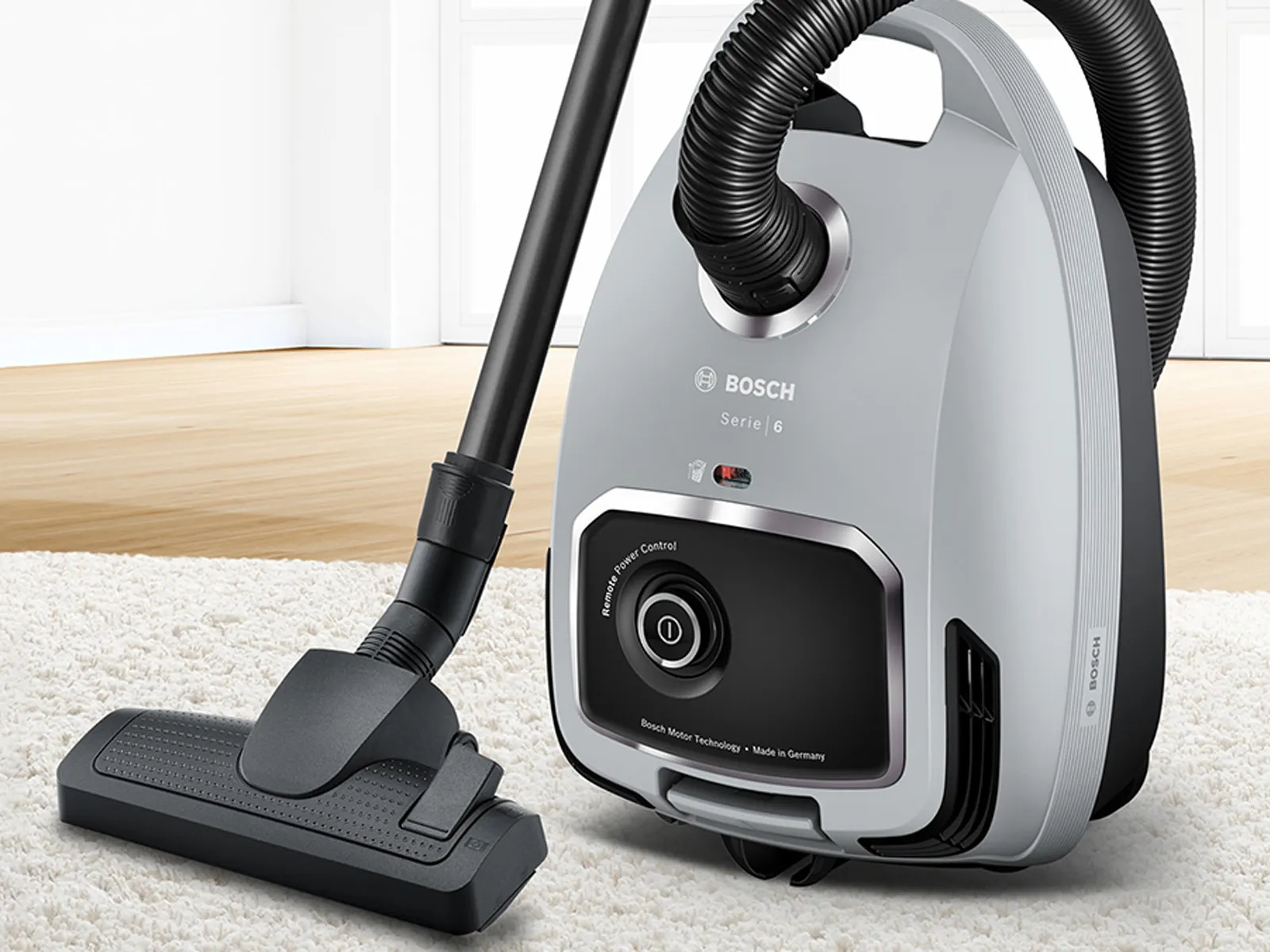 A compact grey cylinder vacuum on a white rug with floor-length windows in the background.