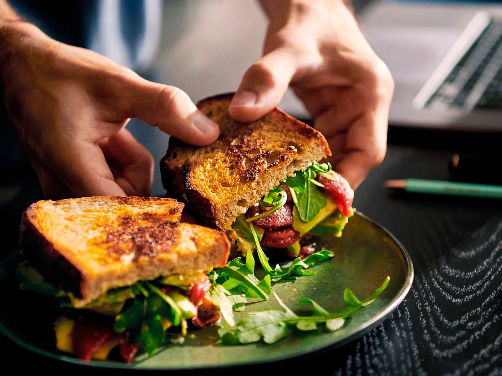 Hænder holder en nygrillet sandwich med sprødt brød, grønt og lufttørret kød, serveret på en tallerken