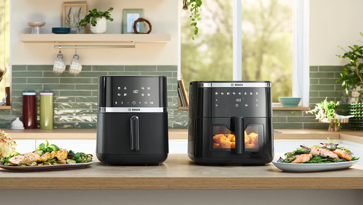 Deux friteuses à air Bosch sont posées sur un plan de travail de cuisine à côté d'assiettes de légumes cuits et de saumon, avec une fenêtre lumineuse en arrière-plan.