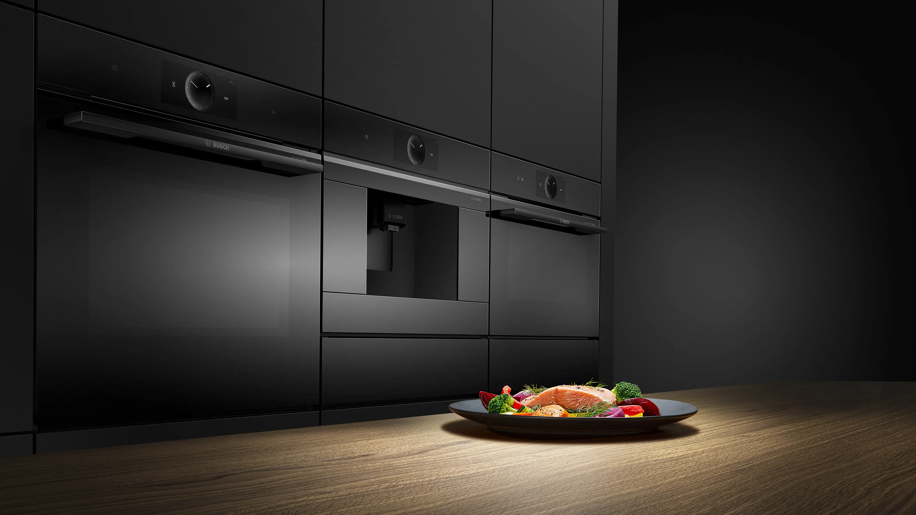 Drei nebeneinander eingebaute, schwarze Bosch accent line Küchengeräte mit einem servierten Lachs- und Gemüsegericht im Vordergrund.