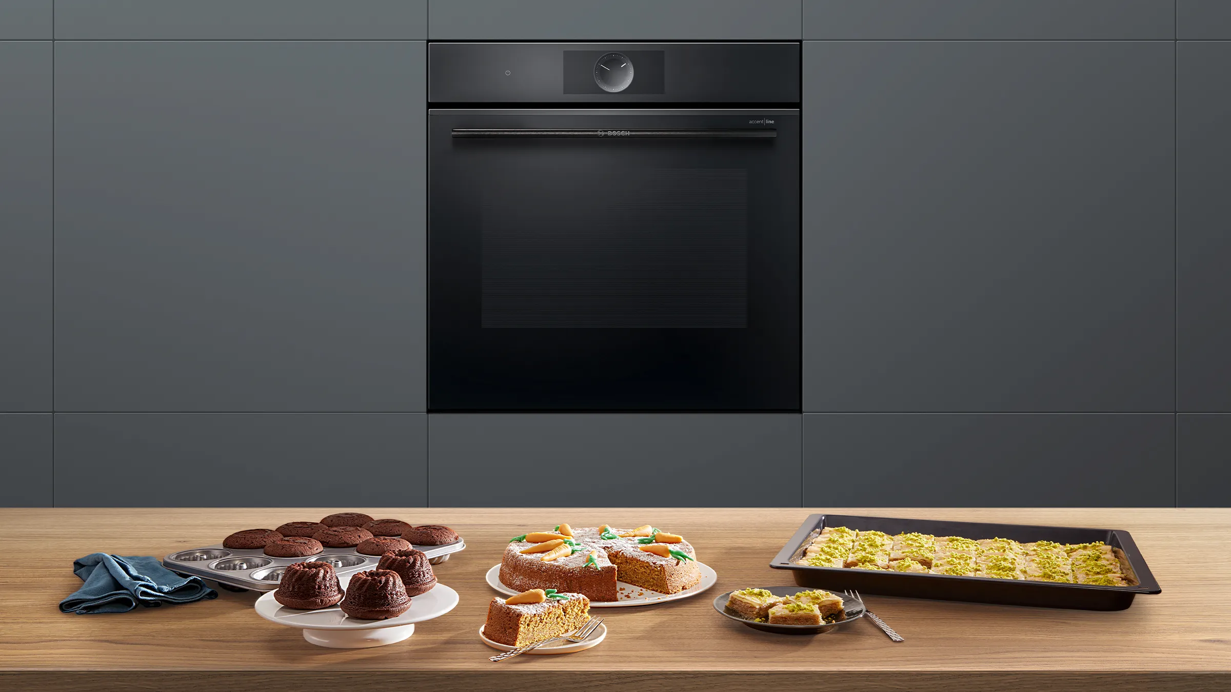 Frontansicht eines Bosch Serie 8 Einbaubackofens in mattem Schwarz mit einer Auswahl an frisch gebackenem Kuchen auf der Holzarbeitsplatte.
