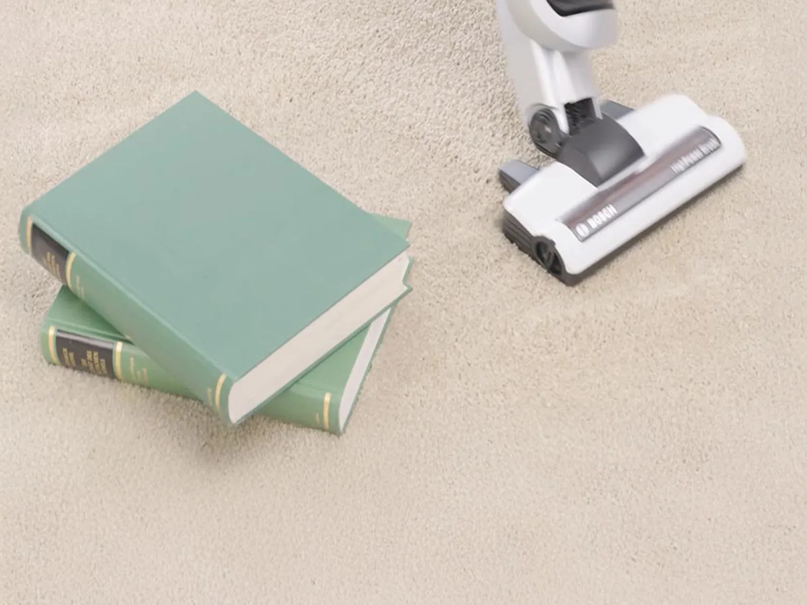 Un aspirateur aspire un tapis. Il y a des livres à côté.