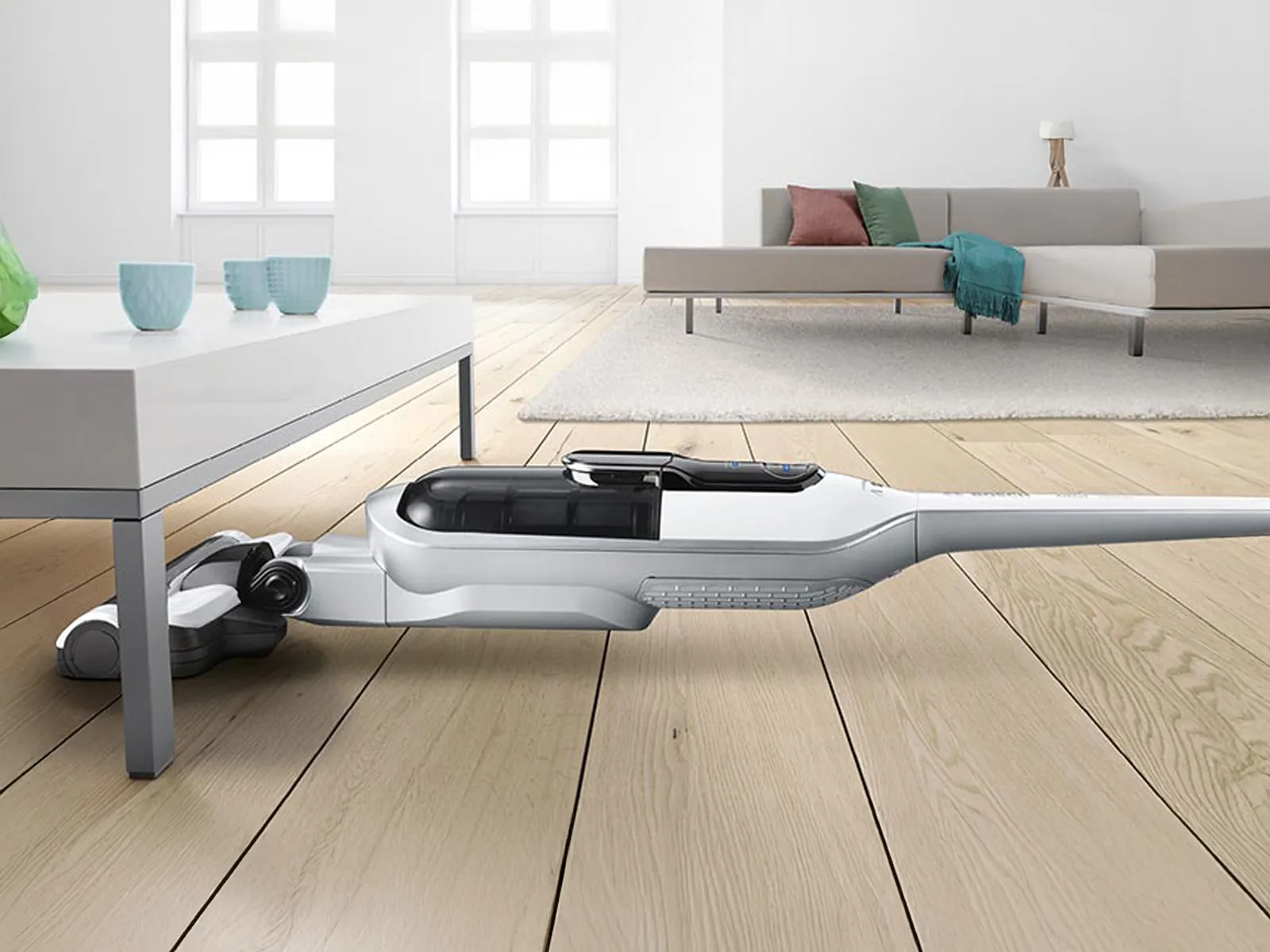 Un aspirateur sans fil nettoie sous une table de salon. Il y a un canapé en arrière-plan.