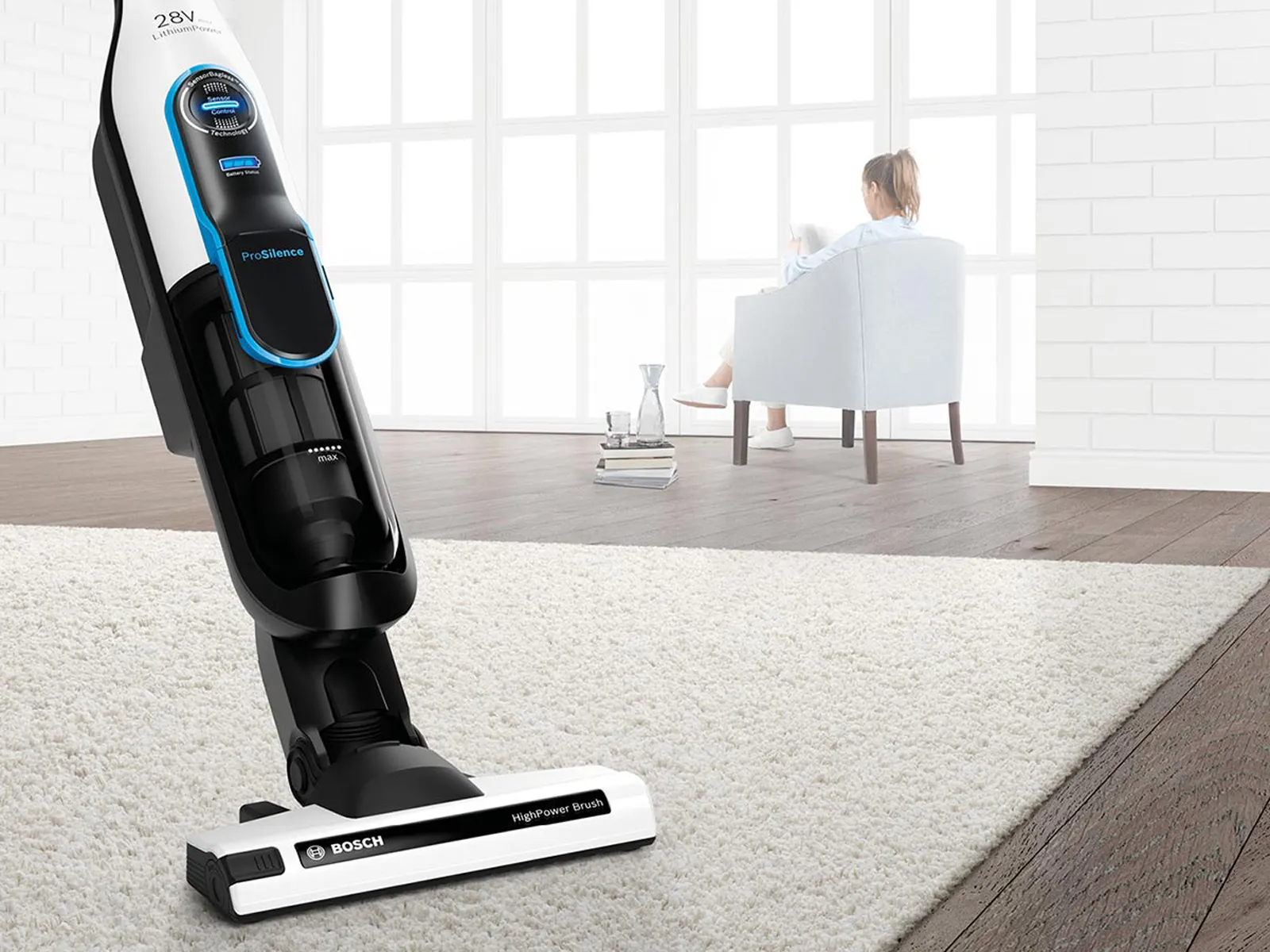 A woman sits in an armchair. In the foreground is an extra quiet cordless vacuum cleaner.