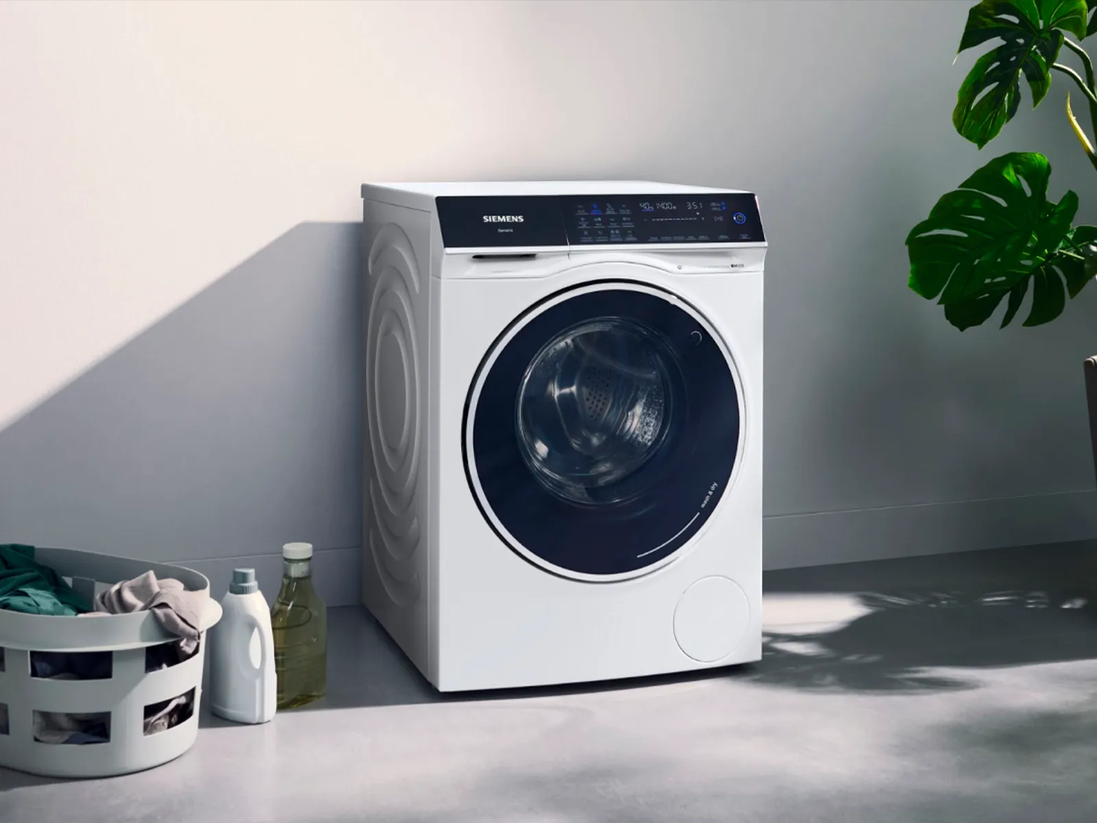 Tvättmaskin med torktumlare i ett modernt rum, till vänster tvättkorg och till höger en planta.