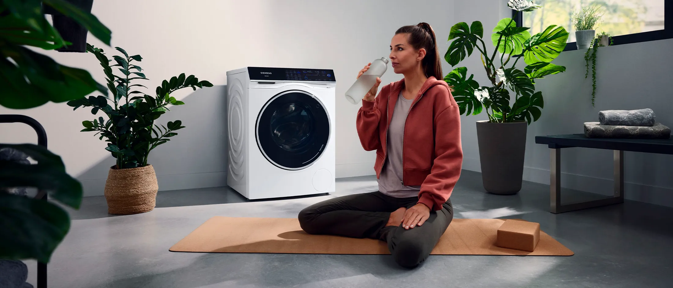 Person sitting cross-legged on a yoga mat, drinking water, next to a washing machine in a modern room.