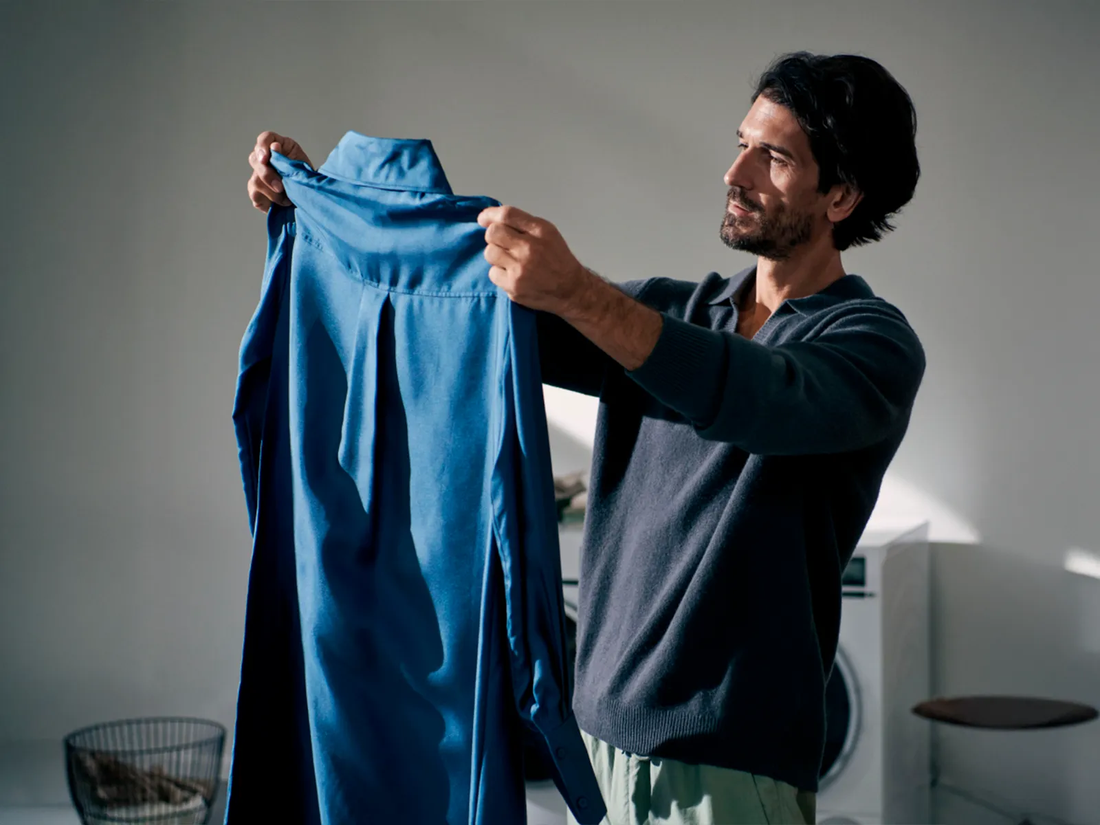 Pessoa a segurar uma camisa azul com um cesto de roupa e a máquina de lavar ao fundo.