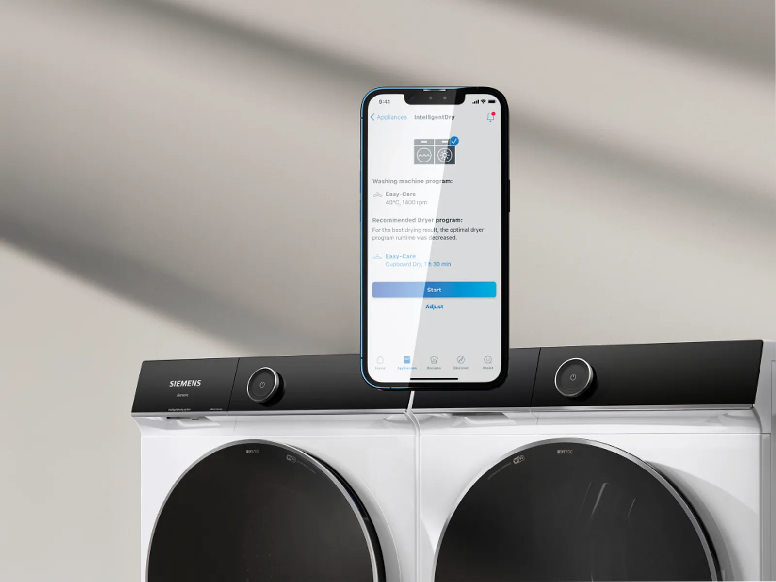 En smartphone som visar en tvättapp placerad på en tvättmaskin från Siemens med en matchande torktumlare bredvid.