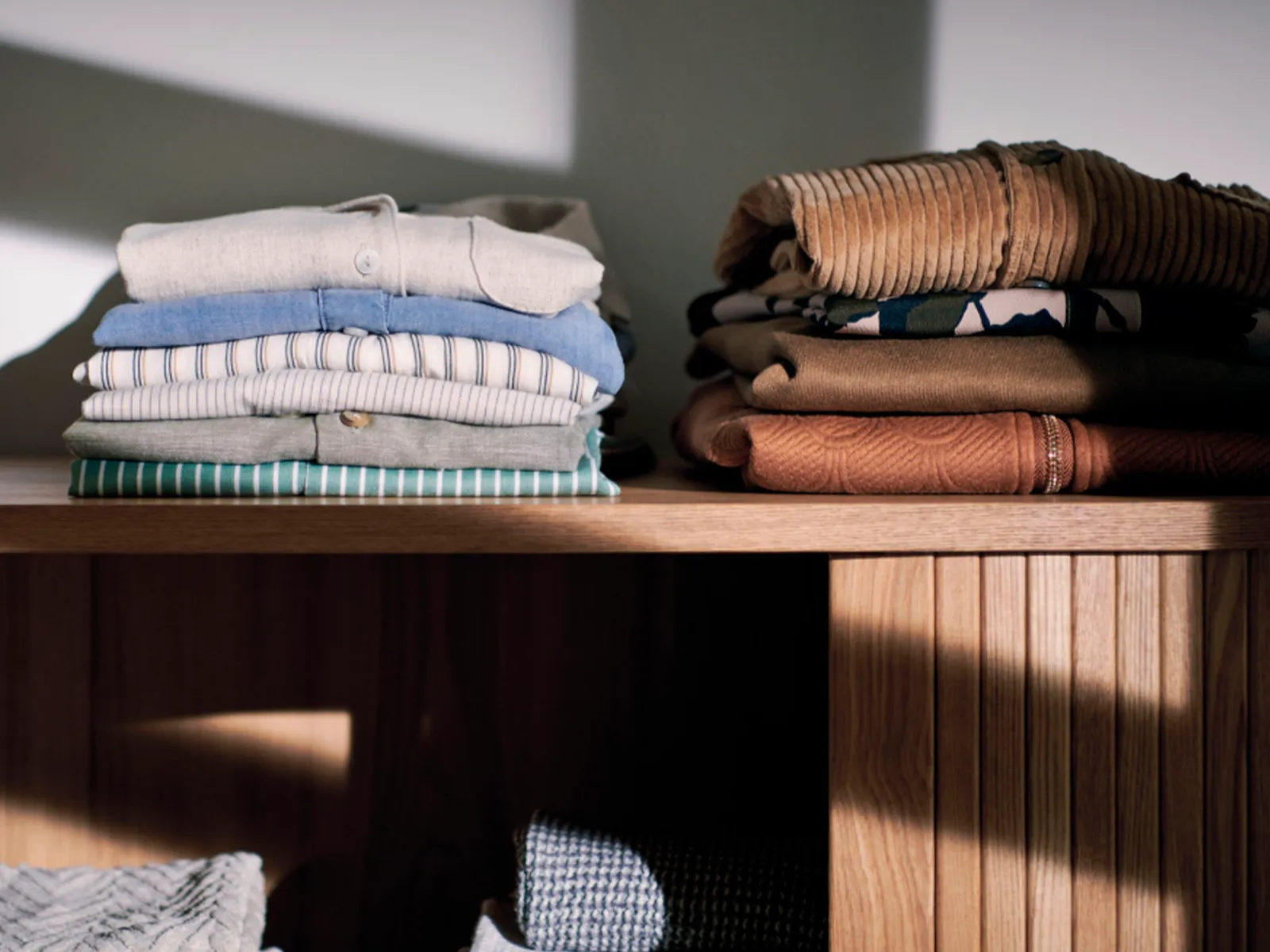 Pilhas de roupas dobradas de forma organizada numa prateleira de madeira sob uma luz suave.