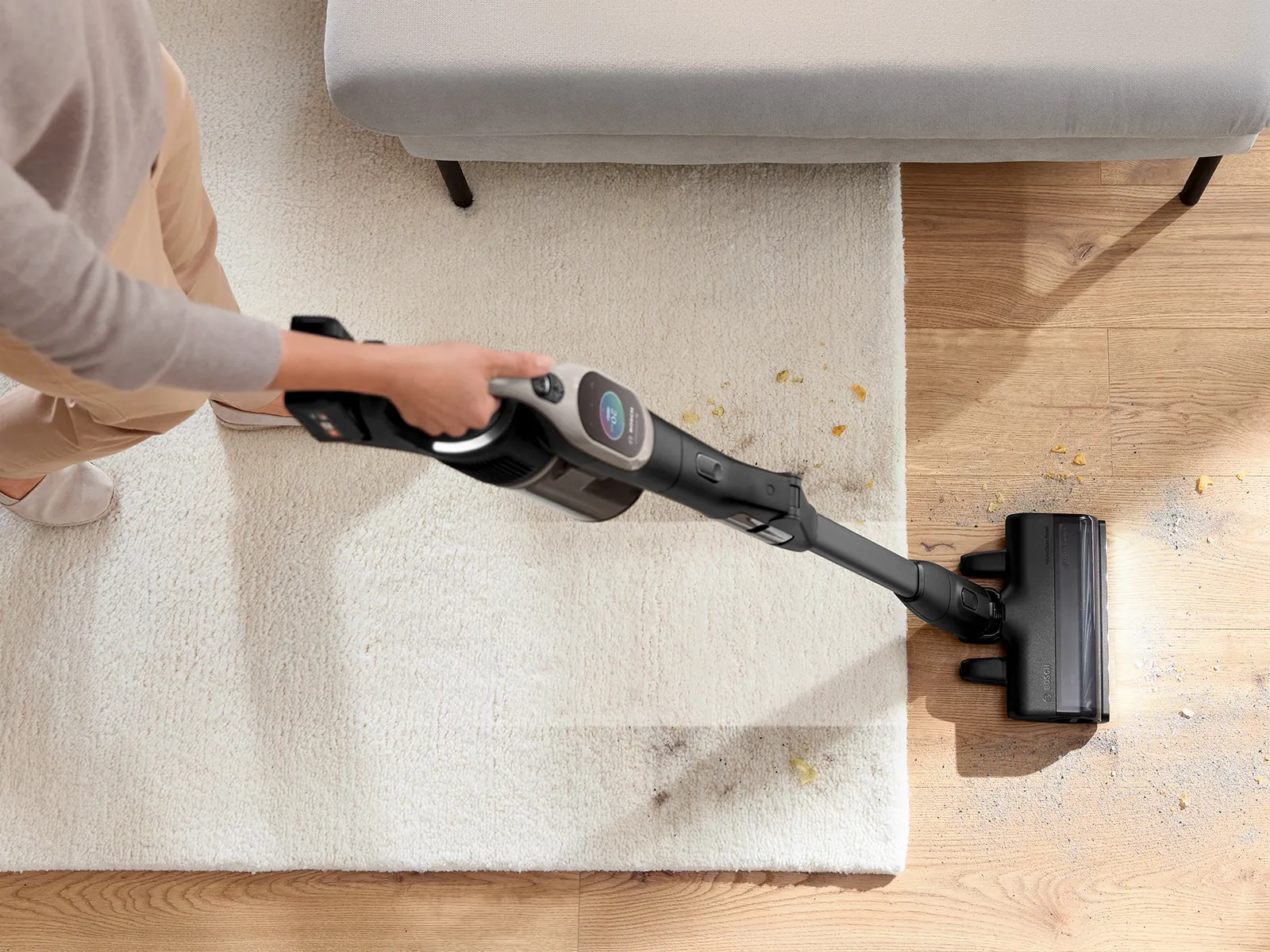 An overhead view of a person using a black cordless stick vacuum cleaner to clean up crumbs on a light-colored rug and a light wood floor. The vacuum's nozzle has a bright LED light illuminating the debris.