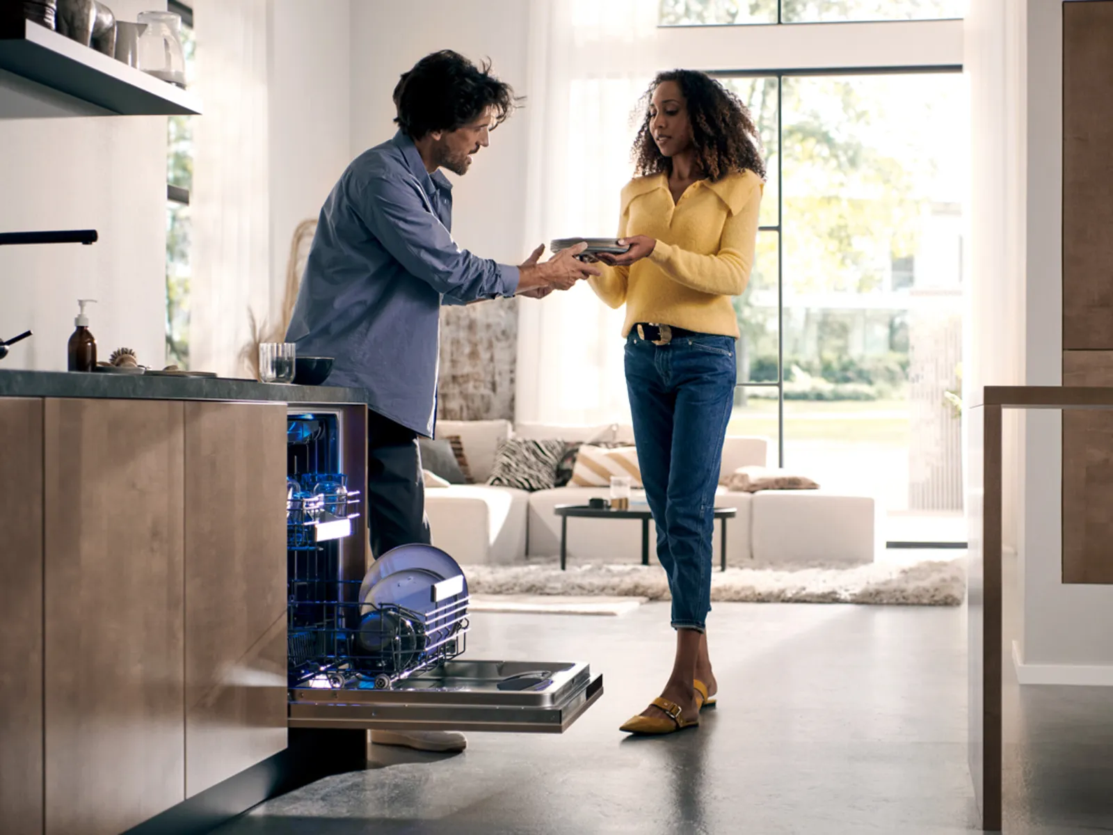 En mand rækker en ren tallerken fra opvaskemaskinen til en kvinde, i et moderne åbent køkkendesign.