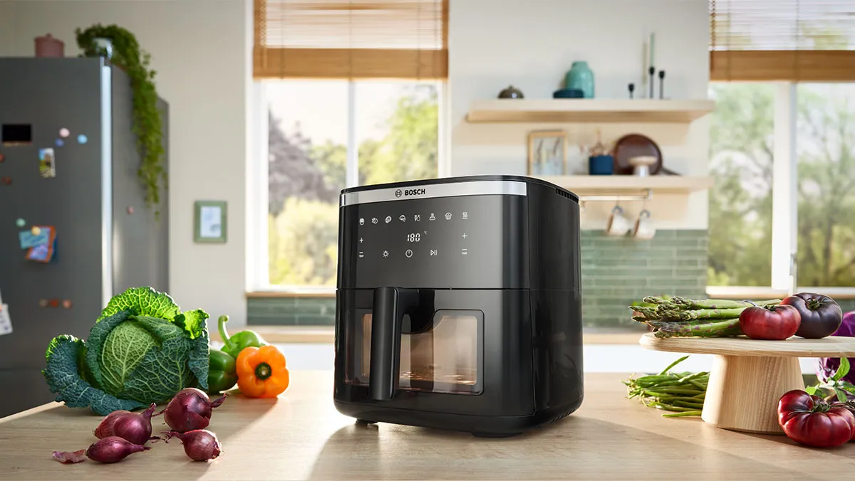 Freidora de aire Bosch negra sobre la encimera de la cocina, rodeada de verduras frescas como repollo, pimientos, cebollas y tomates.