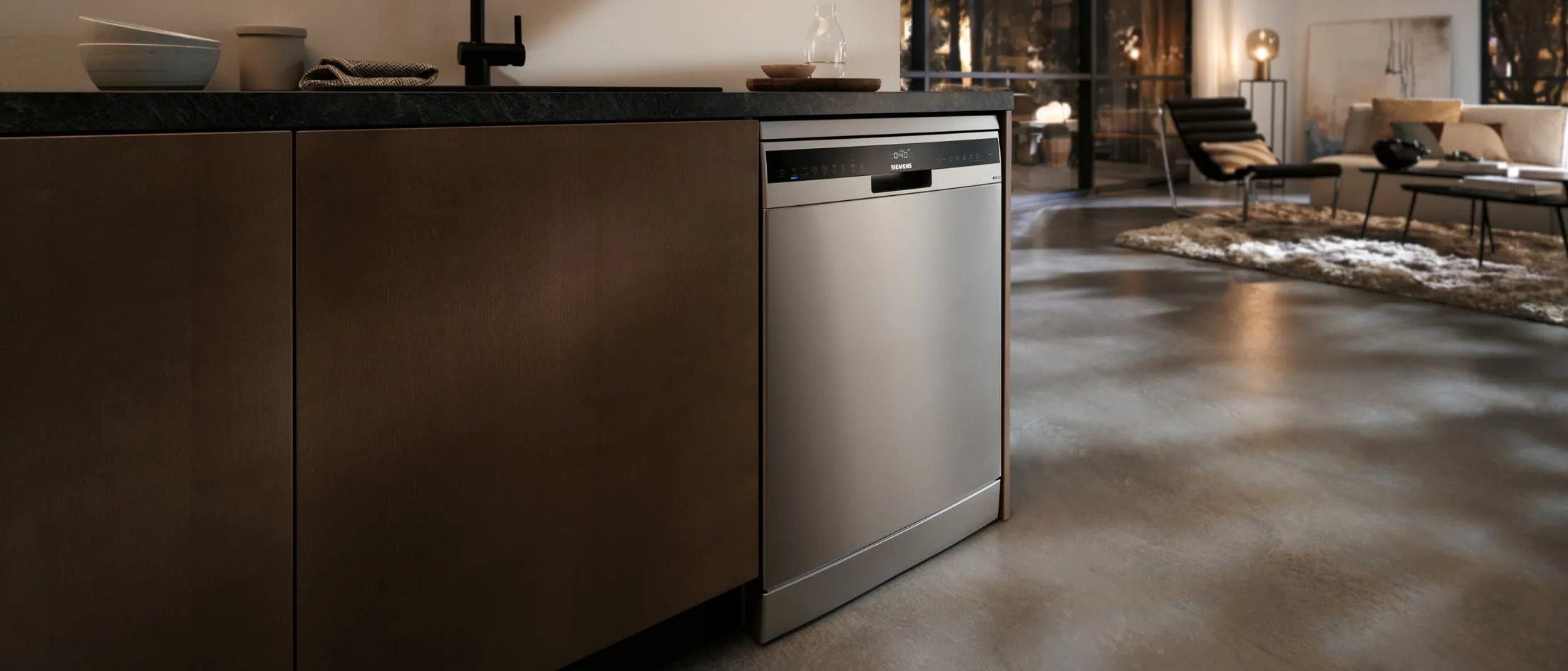 Uma máquina de lavar loiça em aço inoxidável elegante, integrada numa cozinha moderna, com um balcão escuro e um espaço de living aberto e estiloso ao fundo.