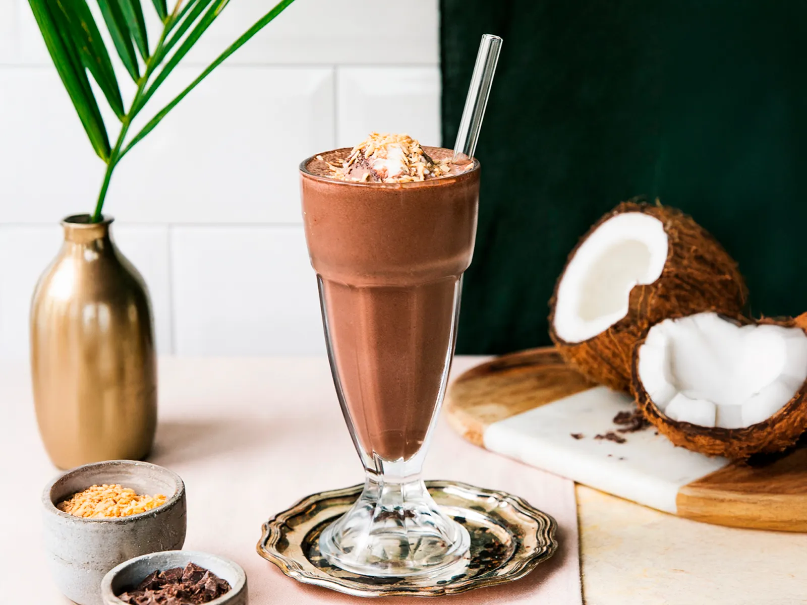 Visoka čaša kremastog čokoladnog smoothieja ukrašena tostiranim kokosom, okružena kokosom i kakaovim zrncima.