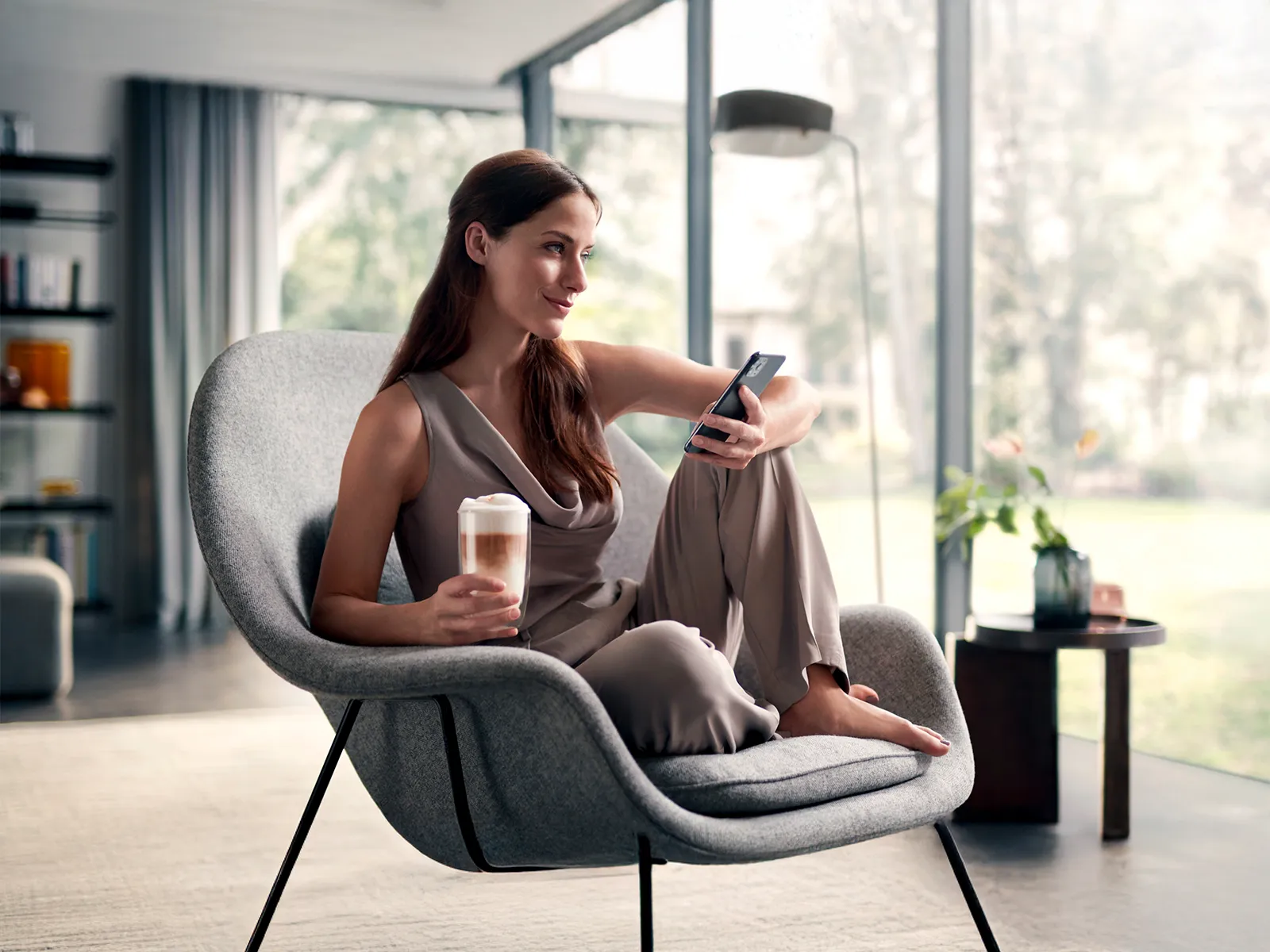 En kvinne som sitter på en stol og holder en smarttelefon og en latte i et moderne stue.