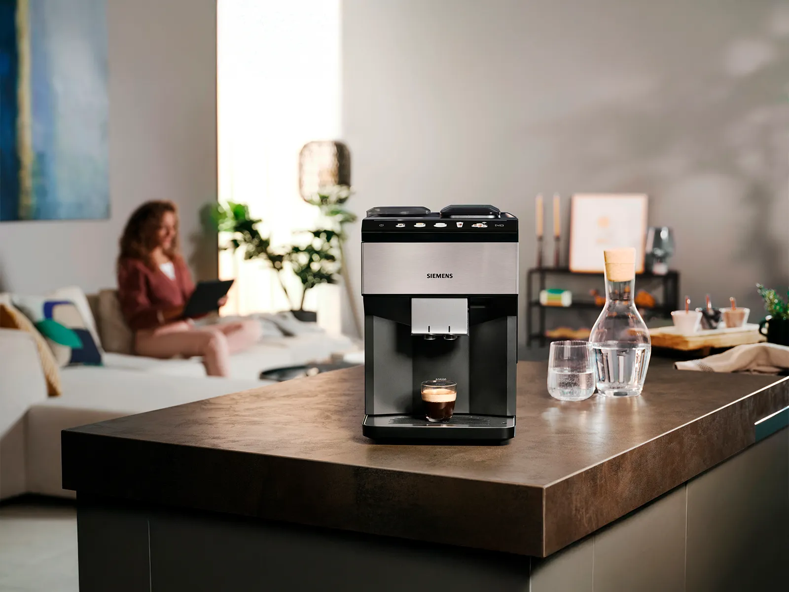 Im Vordergrund steht der EQ500 auf einer Kücheninsel; im Hintergrund sitzt eine Frau auf dem Sofa mit einem Tablet.