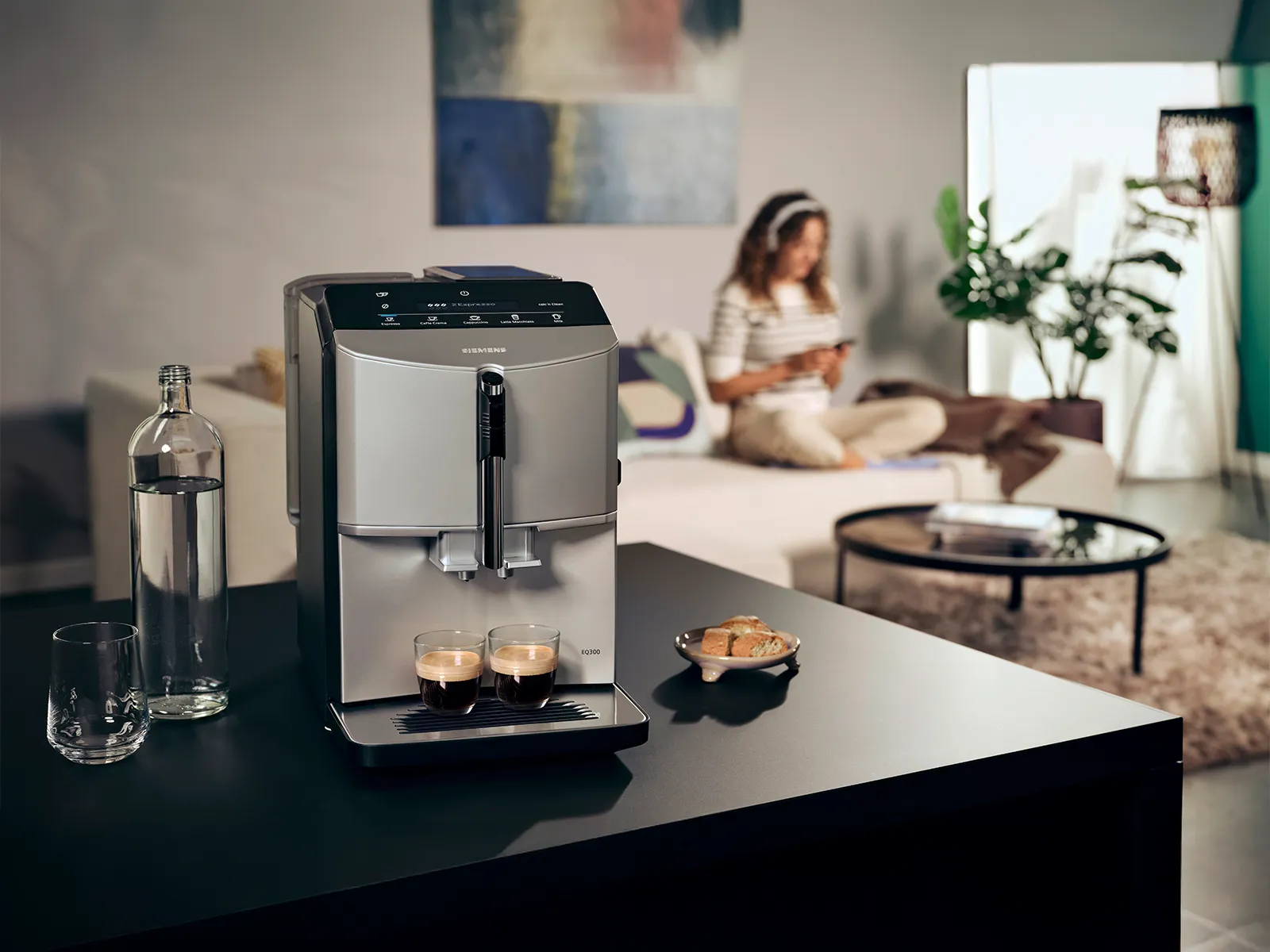 EQ300 fuldautomatisk espresso-/kaffemaskine på en køkkenø tilbereder to espressoer, i baggrunden sidder en kvinde på en sofa.